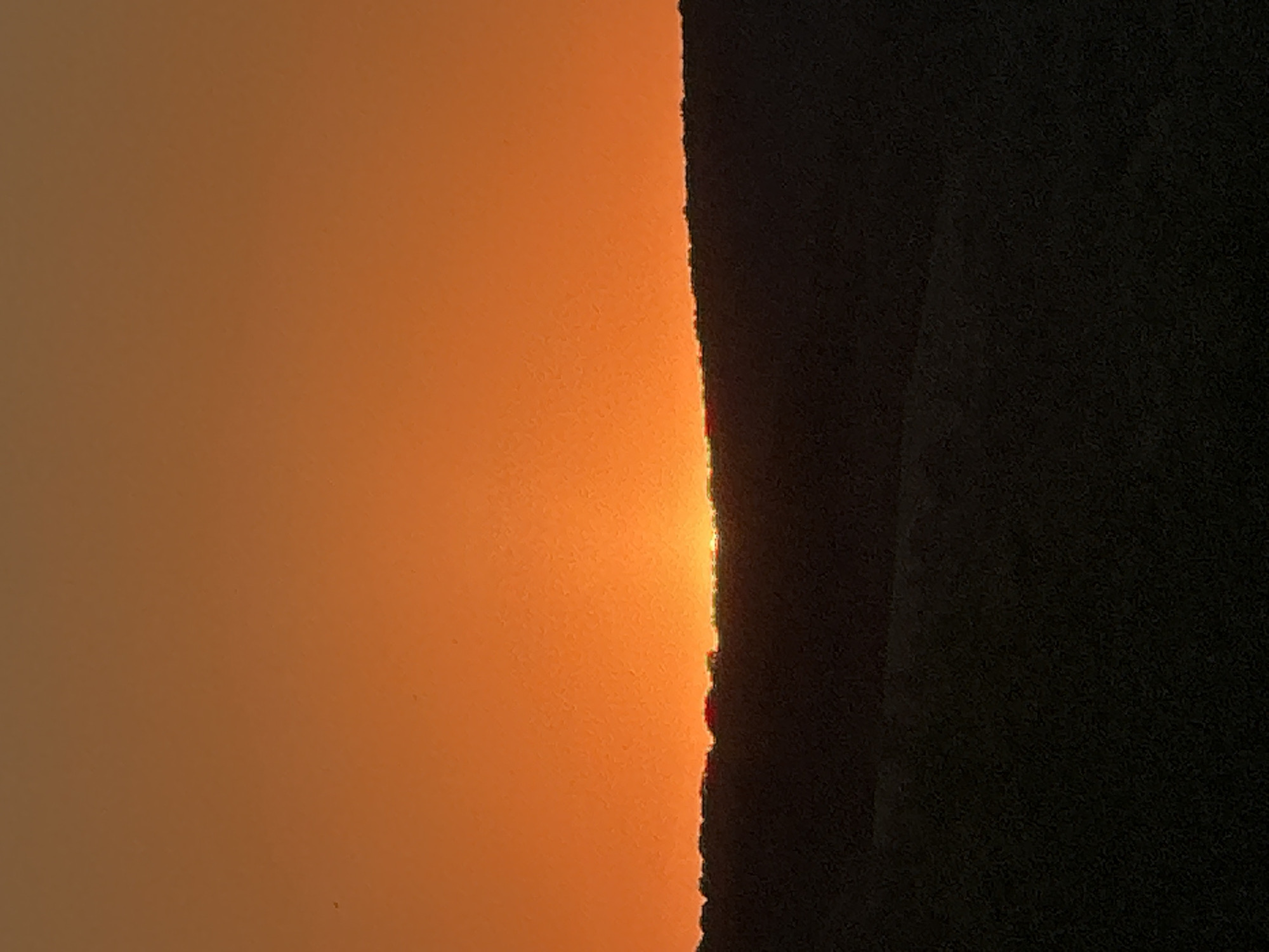 The image captures a sunset over a hilly landscape. The sky is a vibrant orange, gradually fading to a lighter shade towards the top. A dark silhouette of a hill or ridge runs across the lower portion of the frame, with the sun appearing as a small, bright sliver just above the horizon, peeking through a gap in the silhouette. The foreground is a darker silhouette of rolling hills. The scene is devoid of any human presence or man-made structures, depicting a natural landscape at dusk. The location in Makiv, Ukraine, is contextual information not directly derivable from the image itself.