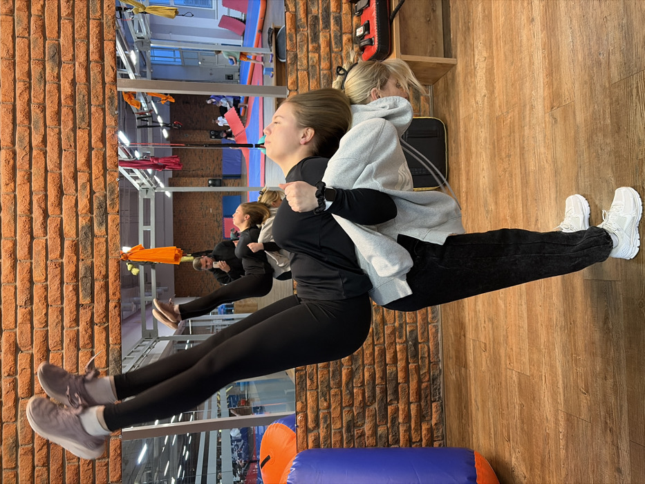 Two females are depicted performing a partner acrobatic or stretching pose inside a gymnasium located in Chaikovsky, Russia. The individual acting as the base is a female, seen in profile, wearing dark trousers, a light grey hooded sweatshirt over a black long-sleeved top, and white athletic shoes. Her upper body is bent forward at the waist, with her head lowered and hands clasped behind her neck, providing support. The second individual, a female, is positioned horizontally across the base's back. She wears black athletic leggings and a black long-sleeved top, with her legs extended vertically upwards, displaying light-colored athletic shoes. Her head is tilted backward, facing upwards.
The immediate environment consists of a light wood plank floor and a reddish-brown brick wall partially visible on the left. A large wall-mounted mirror in the background reflects the broader gym area. The reflection shows various fitness equipment, including red, orange, and yellow aerial silks hanging from the ceiling, blue and red floor mats, black exercise mats, and metal structural components. Several other individuals are visible in the reflection, including a male in a dark top holding a smartphone, seemingly capturing the scene, and other adults and children in the distant background of the reflected space. On the far right, a black and red striking pad rests on a shelf, and a blue and orange inflatable cylinder is partially visible on the far left.