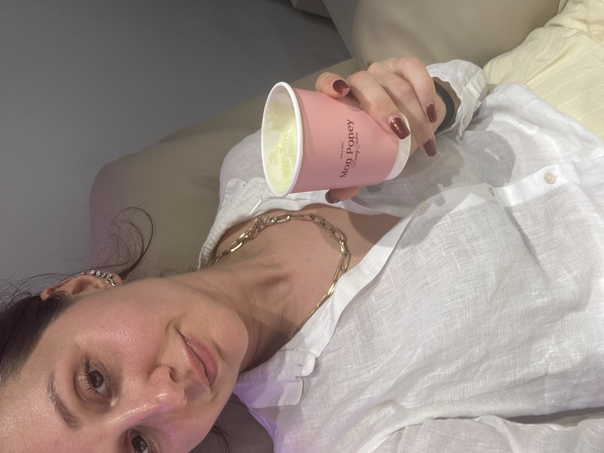 A woman reclines on a neutral-toned, soft surface, likely a sofa, while holding a pink cup with the branding "Mon Poney Beauty Salon Since 2017" on it. The cup contains a light-colored beverage, possibly matcha. The woman wears a white linen shirt, partially unbuttoned, revealing a gold chain necklace around her neck. Her nails are painted a dark, glossy shade. The background includes a light gray wall, while the woman's left ear has multiple piercings. There's no immediate evidence to confirm or deny the location is Dubai, United Arab Emirates.