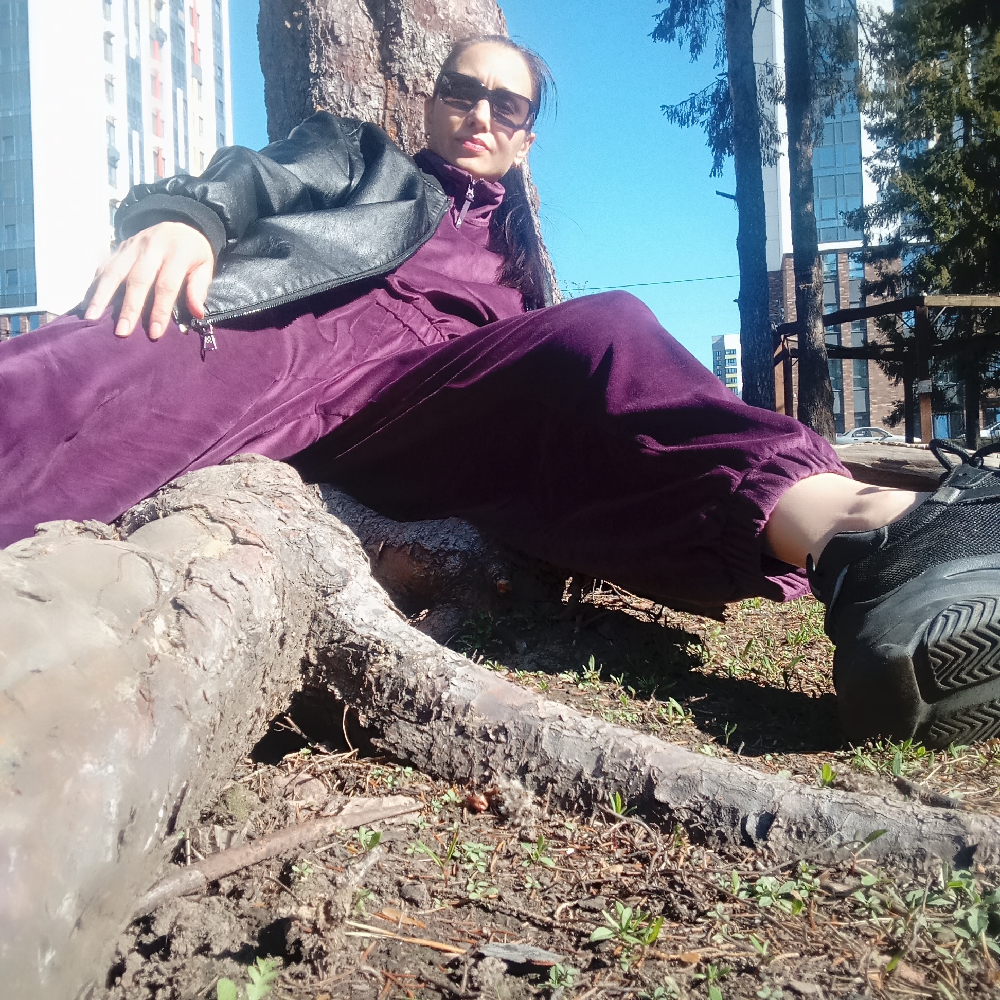 A woman in a dark jacket and purple jumpsuit sits on the exposed roots of a large tree. She wears sunglasses and looks towards the camera. Her left hand rests on her leg, while her right leg is extended towards the camera, with a black shoe prominently displayed. The background includes buildings, trees, and a blue sky. The location is Izhevsk, Russia. The ground is covered with soil, small plants, and fallen pine needles.