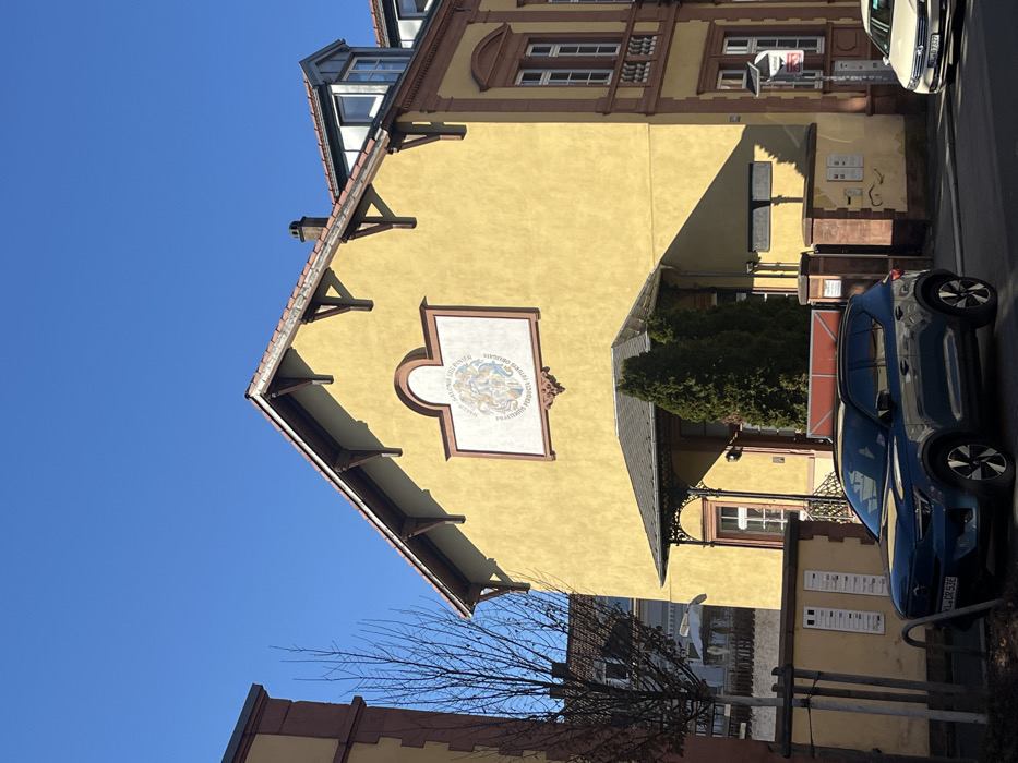 The image depicts a two-story building, identified as the Corps Palatia-Guestphalia, located at Kanalstraße 13, Kaiserslautern, Germany, under a clear blue sky. The building exhibits a warm yellow/ochre stucco facade accented with reddish-brown trim around its windows, cornices, and roofline. Its pitched roof is covered with reddish-brown tiles and features exposed wooden corbels supporting the eaves, along with a visible chimney stack and a dormer window on the right side.
A large, rectangular decorative crest, framed by a reddish-brown arch, is centered on the upper facade. The crest contains intricate artwork and the Latin inscription "COLLEGIVM FALCONIS SED POSTEA FRATERNITAS PERDOCTA FUTURA PEREGRINA". Below this crest, a recessed main entrance is covered by an ornate, dark metal canopy. Two tall, conical evergreen shrubs flank the entrance.
In the foreground, a dark blue Volvo XC40 Recharge SUV (license plate KL DR 53E) is parked on the asphalt street directly in front of the building. Further down the street on the right, a white sedan (license plate KL D 7327) is partially visible. Metal bollards or bicycle racks are positioned on the sidewalk near the blue SUV. To the left of the building's entrance, a low wall displays several white notices. A bare deciduous tree stands to the left, partially obscuring an adjacent building of similar architectural style. Strong natural sunlight casts distinct shadows across the scene. No individuals are visible.