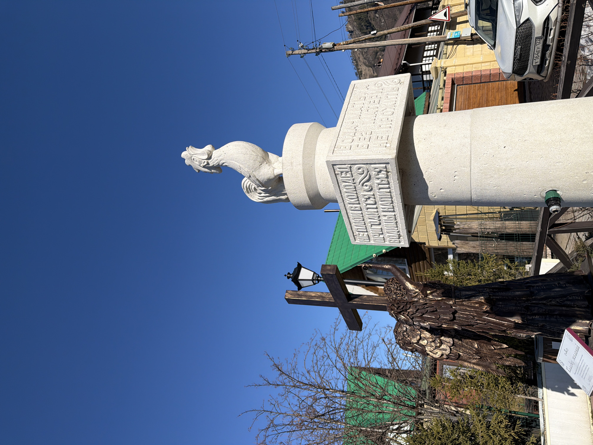 Here is a description of the image:
The image shows a monument in the city of Manzherok, Russia, featuring a white statue of a rooster atop a stone pillar inscribed with text in Russian. Below the rooster monument is a wooden sculpture of an angel attached to a cross with a lamppost. A sign with more Russian text and the price "4000 rub" sits next to the angel. The background includes buildings, power lines, trees, a blue sky, and a portion of a white car with the license plate "K164EY 122".