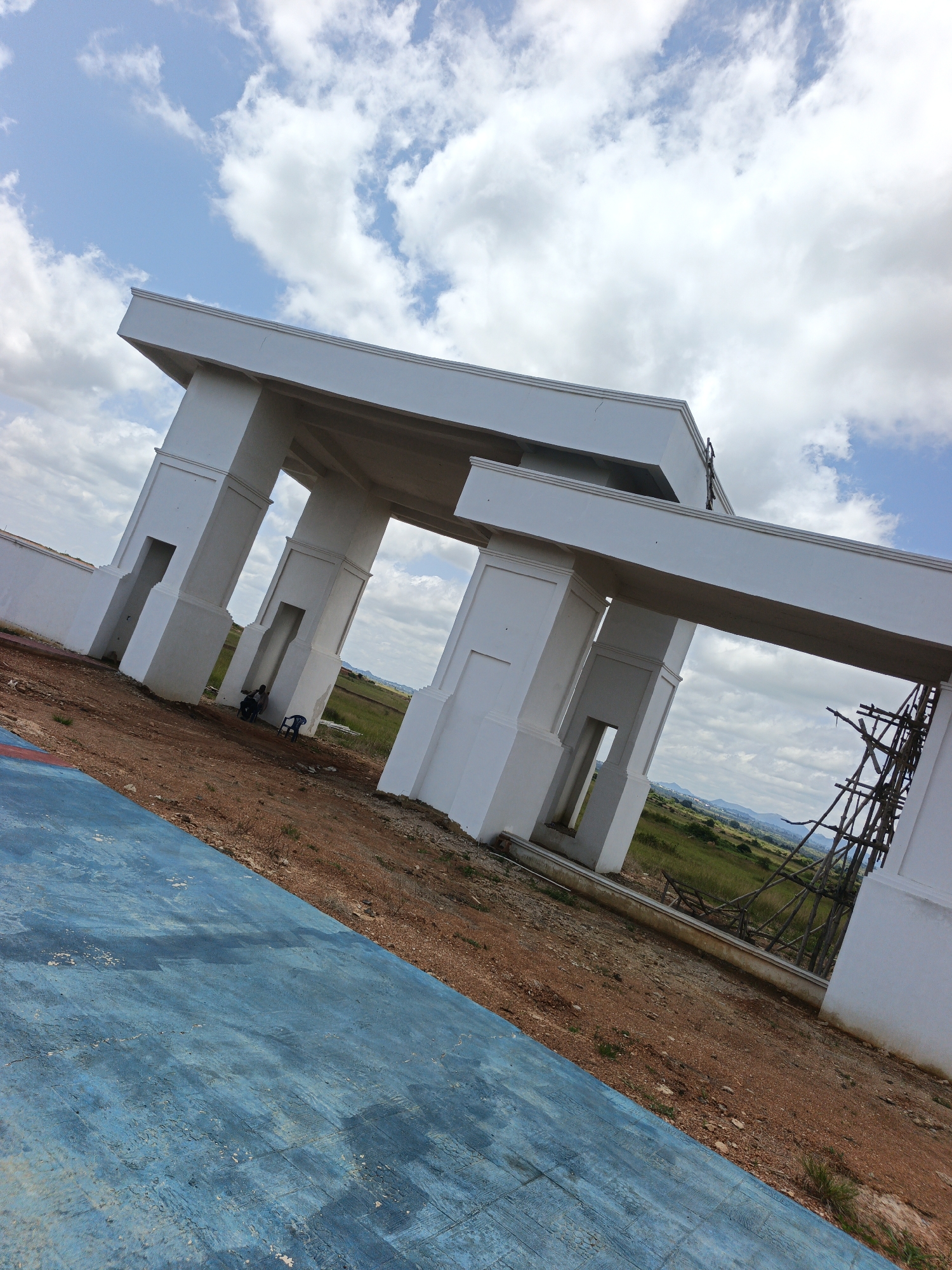 The image shows an unfinished white structure, possibly an entrance gate, against a cloudy sky. The structure features large columns and horizontal beams, with open spaces between the columns. In the foreground, a blue-painted surface slopes diagonally. The ground around the structure is dirt and sparse vegetation. In the background, there is a green field with some trees and hills in the distance. Construction scaffolding is visible to the right of the structure. Two individuals are seated under the structure, one on a blue chair and the other on the ground. The location is identified as Du, Nigeria.