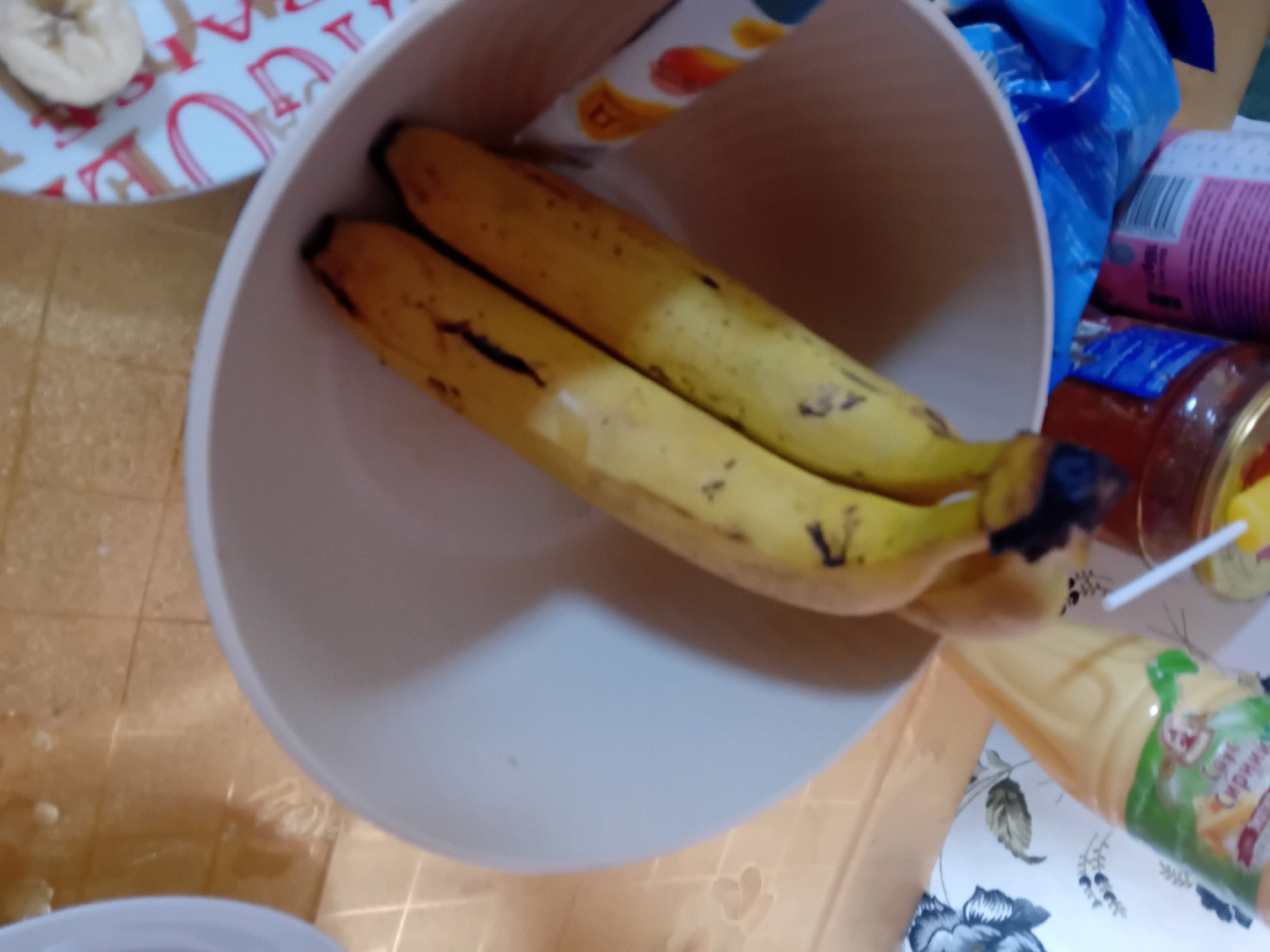 Two bananas are in a white bowl. The bananas are yellow with some brown spots. In the background, there is a jar of jam and a blue plastic bag. There is also a plate with a sliced banana on it. The scene is on a table with a patterned tablecloth. The location is not specified.