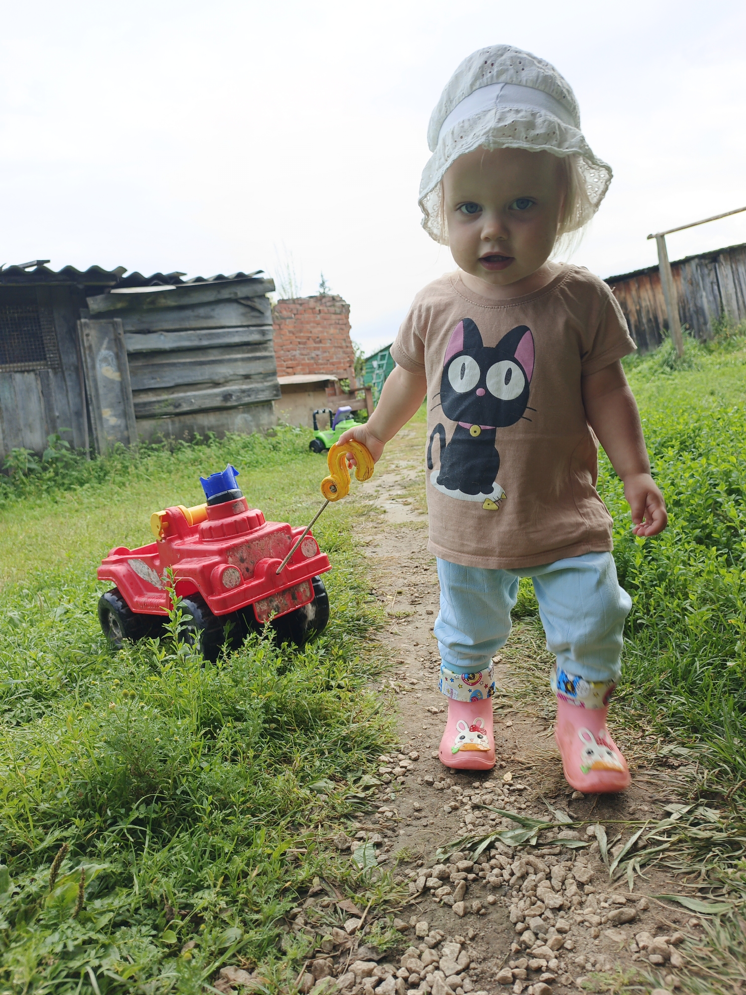 A young Caucasian girl, approximately 2-3 years old, stands on a gravel path in a rural outdoor setting that is identified as Medvedyovka, Russia. She is wearing a white eyelet sun hat, a light brown t-shirt with a black cat graphic, light blue pants, and pink rain boots adorned with rabbit images. The girl is holding a yellow tow hook attached to a red plastic toy truck. The truck has a blue and yellow plastic piece on top and appears to be made of durable plastic. The background features a wooden structure, possibly a shed or fence, overgrown with green grass and weeds. The sky is overcast.