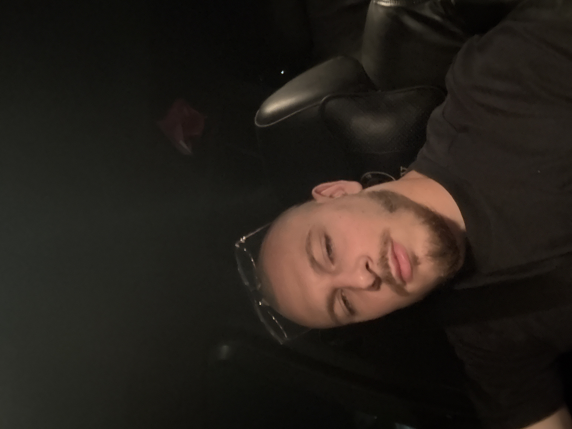 A selfie captures a young man from the chest up, seated inside a car at night. He has a shaved head, a goatee, and is wearing a black t-shirt. A pair of glasses rests on top of his head. The car's interior is dark, with a glimpse of a black leather seat and headrest visible. A small, blurry red object is seen in the upper right corner of the frame. The scene is entirely indoors and does not provide any information to determine the location in the city of Stara Zagora, Bulgaria.