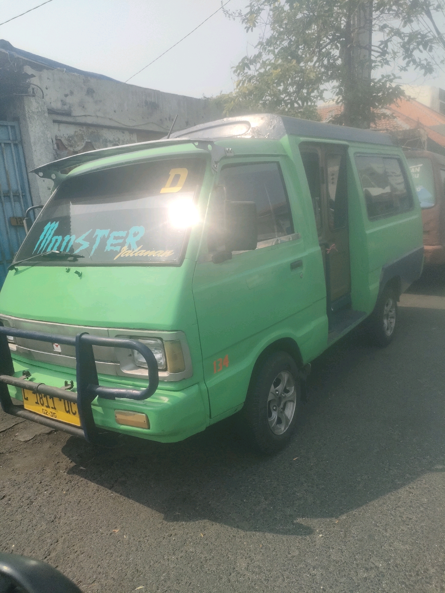 A bright green, slightly weathered van with a dark blue trim is parked on an asphalt road. The van has tinted windows, and a large, dark spoiler is mounted on the roof above the windshield. The windshield itself features two stickers: one in blue that reads "MONSTER" in a stylized font, and another in yellow that reads "Jalanan". A yellow license plate, partially obscured by a dark blue bull bar, displays the text "L 1811 UC" and "02-30". A number "134" is visible in orange on the lower passenger side of the van. The environment appears to be an urban setting in Surabaya, Indonesia, with a building with a gray, weathered facade and a corrugated roof visible in the background. Trees and a utility pole are also present in the background, under a hazy, bright sky. The sunlight creates strong glare on the van's windshield and casts shadows on the road.