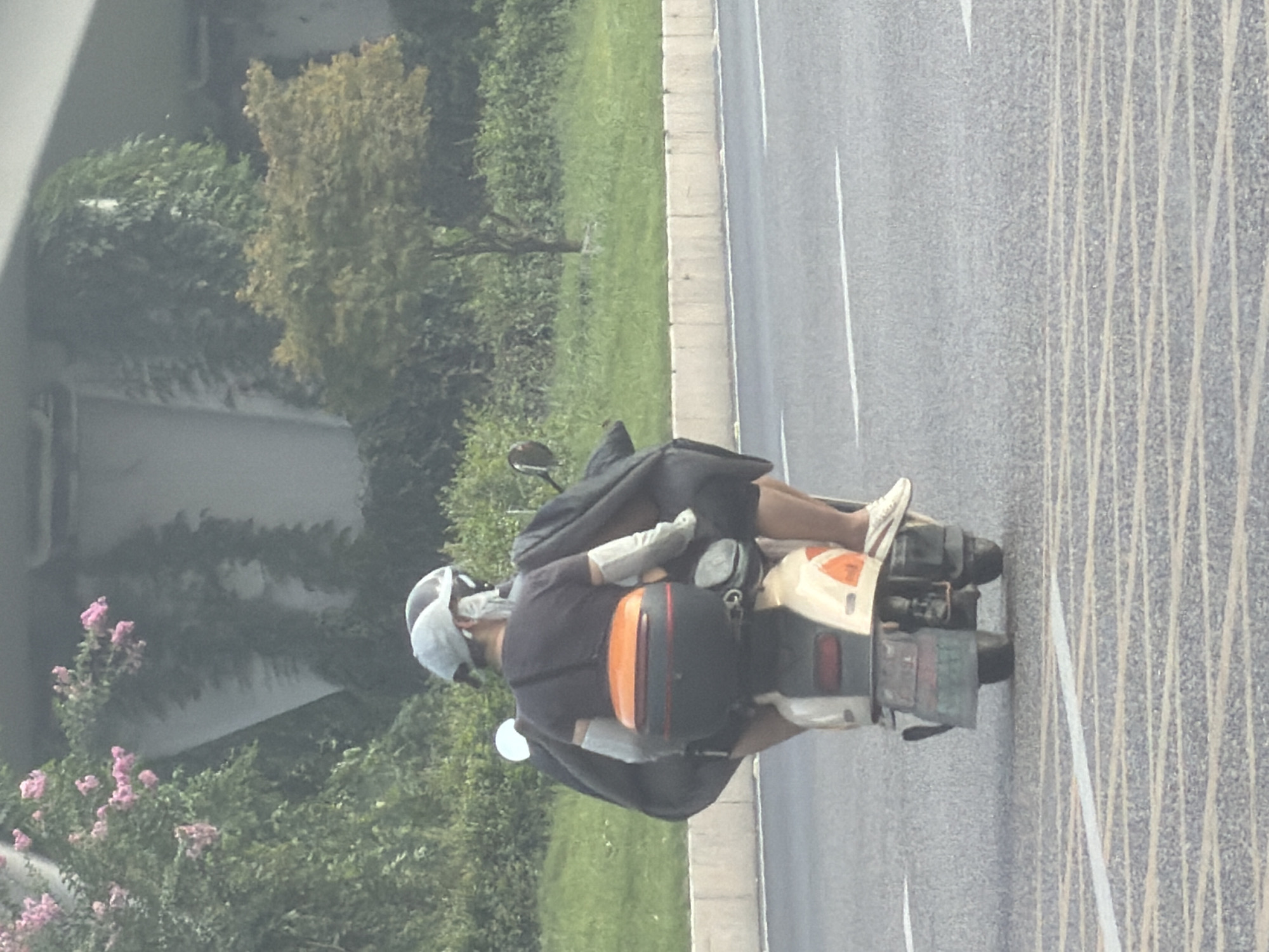 In Jiangjin, China, on a street with a grey asphalt surface marked with white lines, a two-person scooter is in motion. The scooter has a tan and white body with an orange and black storage box on the rear. The person in front wears a grey helmet and a dark t-shirt, with light-colored arm sleeves. The person behind also wears a dark t-shirt, visible legs, and white sneakers with red stripes. Both individuals are covered by dark fabric. The background features green vegetation, including bushes and trees, with a grey overpass structure and pink flowering plants visible on the left.