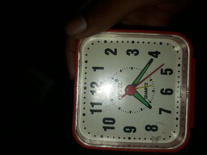 The image shows a red and white alarm clock with black numbers. The clock face reads 5:54. The clock is held by a hand in the lower right-hand corner of the image. The image was likely taken at night, as the background is dark. The location of this image, based on coordinates (latitude: 20.38, longitude: 78.117), is Yavatmal, India.