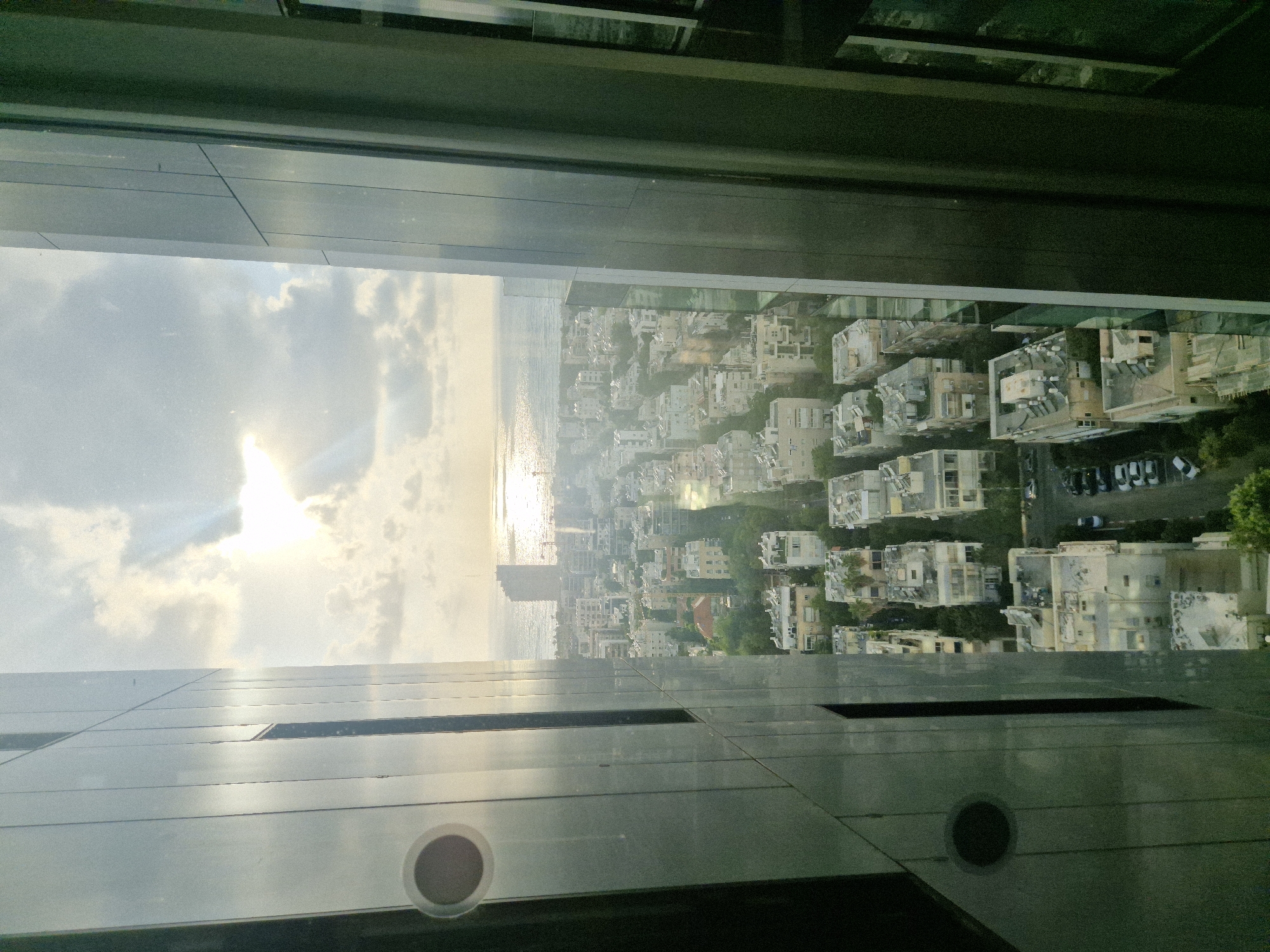 View from a high-rise building in Tel Aviv, Israel, looking toward the Mediterranean Sea. The scene includes a cityscape with closely packed residential buildings, the sea with sunlight reflecting on the water, and the sky with clouds. The foreground shows the architectural features of the building from which the photo is taken, including metallic panels and circular vents. The image is rotated.