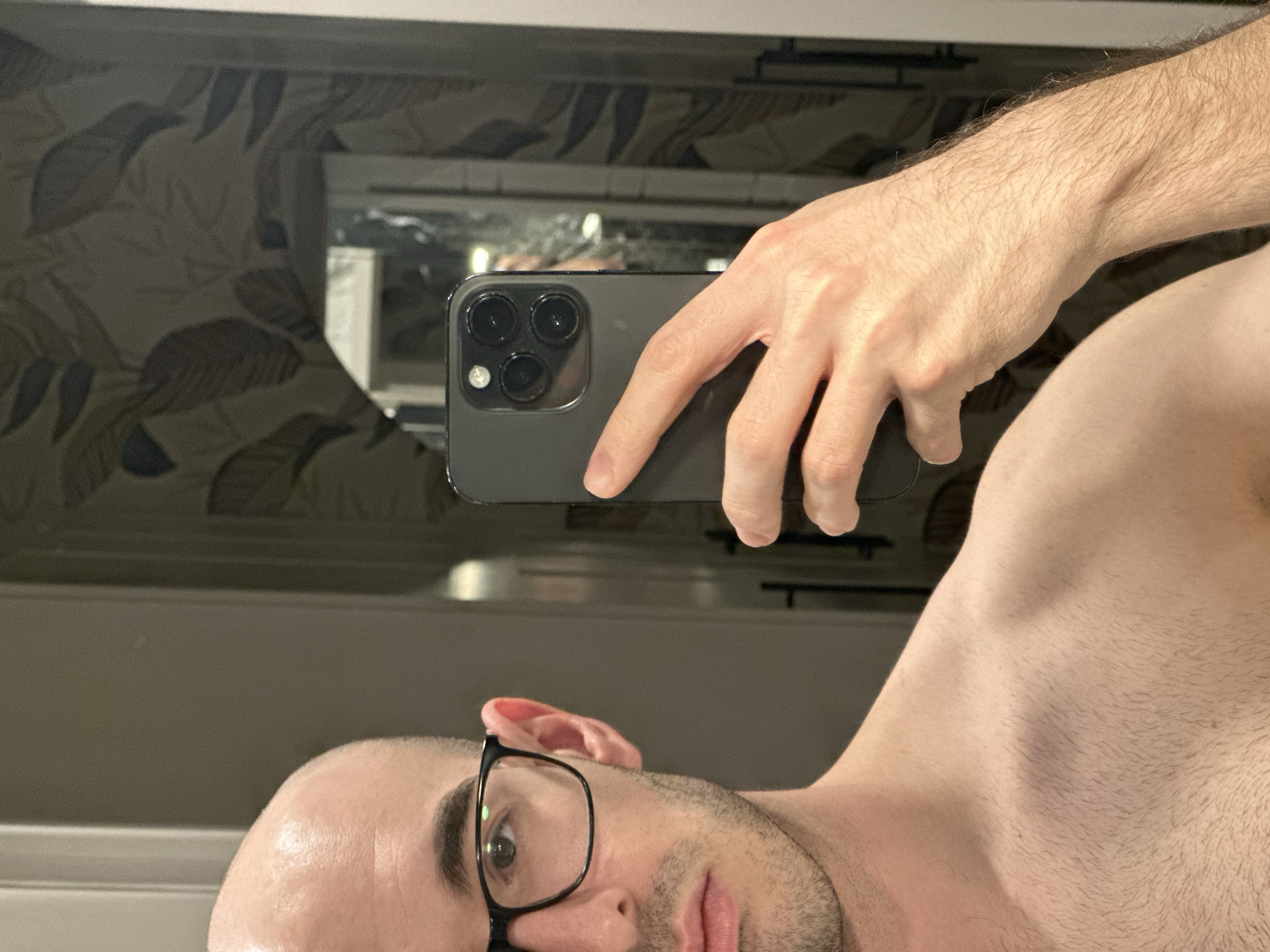 A shirtless person with a shaved head and wearing glasses is taking a selfie in a bathroom. The person is holding a dark-colored smartphone in their right hand, and the camera's lenses are visible. The background includes a mirror with a reflection of the bathroom's wall, which is decorated with a leaf pattern. No evidence indicates the city of Kazan, Russia.