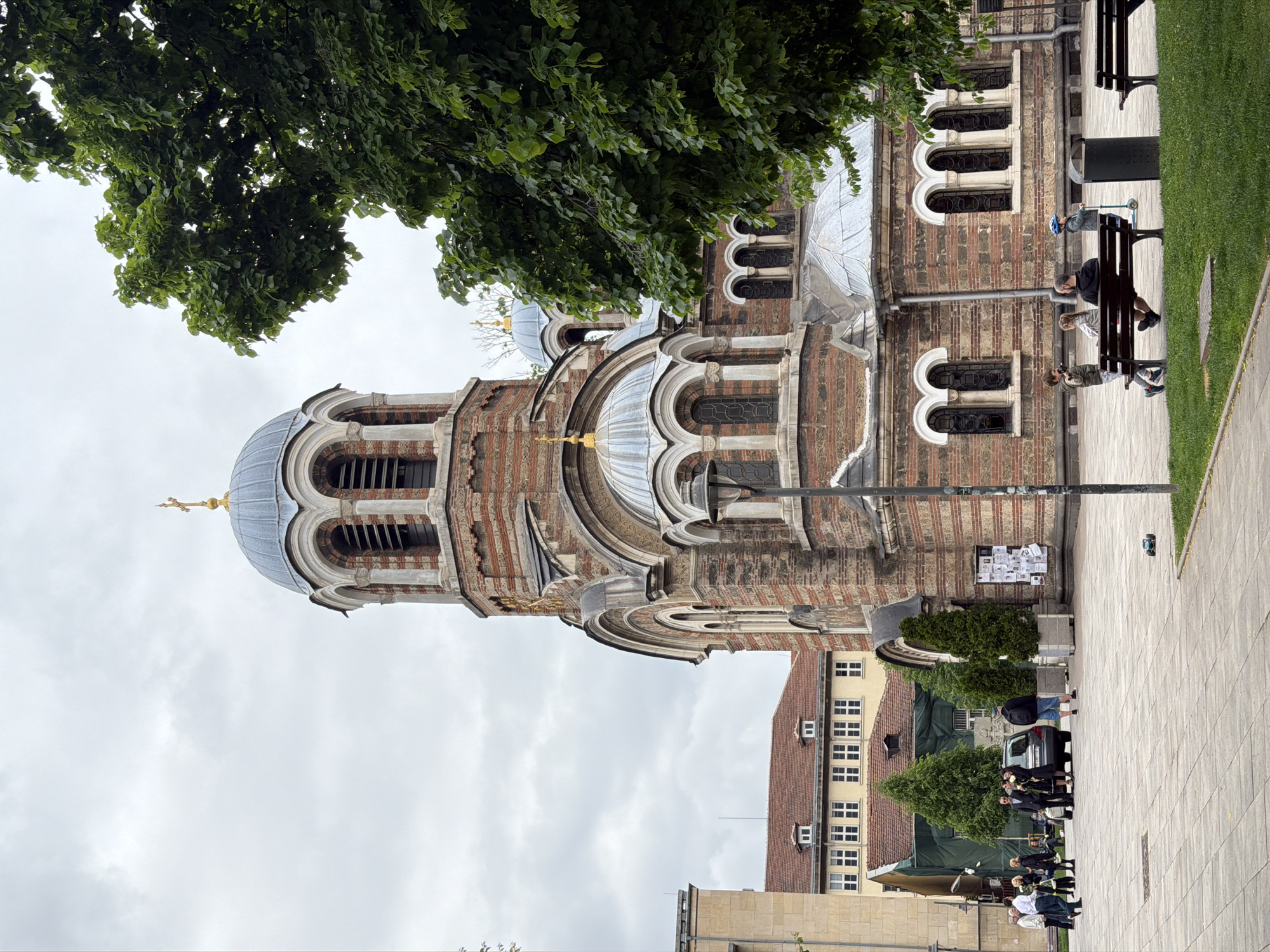 A vertical shot shows a church in Sofia, Bulgaria, on a cloudy day. The church is tall with a domed roof, windows, and a cross on top. It is constructed of alternating layers of brown brick and light-colored stone, in horizontal bands. A tree branch with green leaves occupies the top left corner of the shot. A sidewalk, paved with large square tiles, runs along the right edge of the frame. Several people are walking on the sidewalk, and others are sitting on benches. Behind the church, other buildings with windows and red tile roofs are visible. In the upper right corner of the frame, there is a small patch of green grass.