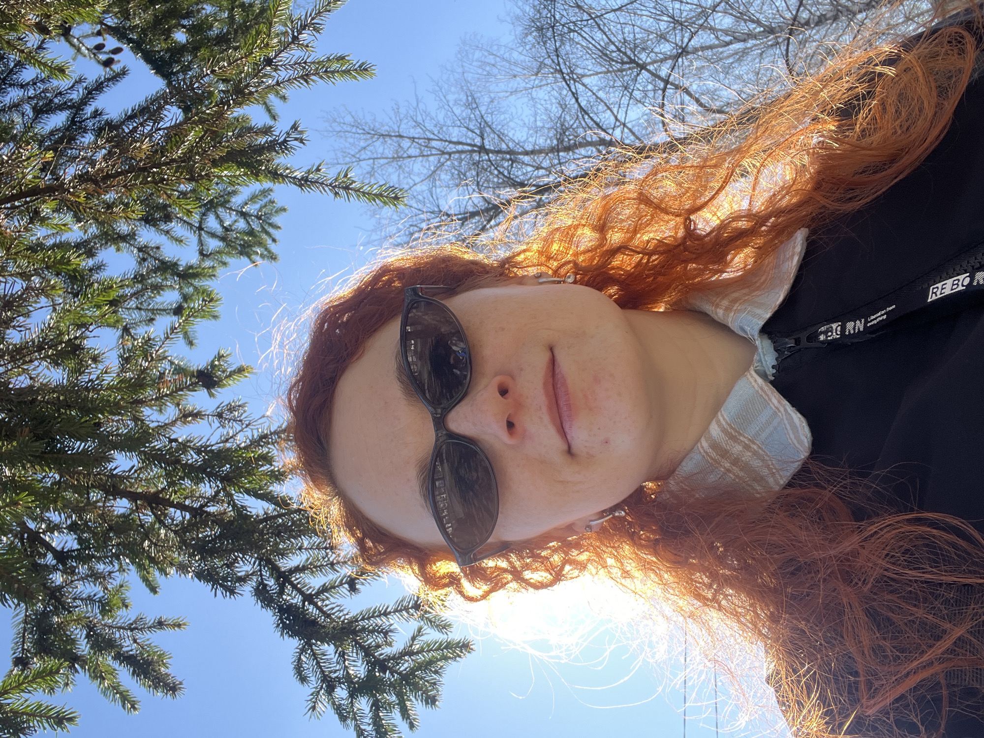 A photograph taken from a low angle depicts a woman with red curly hair wearing sunglasses. She is positioned against a backdrop of a clear blue sky partially obscured by the branches of an evergreen tree to the left and bare tree branches above. The woman is wearing a dark jacket with the word "REBORN" visible on the collar. The sunlight is shining brightly, particularly highlighting the edges of her hair. The location appears to be Chaikovsky, Russia.