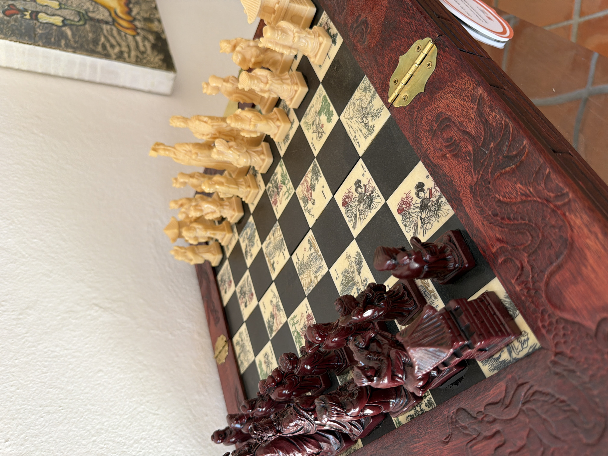 An ornate, Asian-themed chess set is prominently featured, captured from an elevated, angled perspective. The chess board is made of contrasting black and light-colored squares. The light squares are adorned with intricate, hand-drawn illustrations, some with subtle color accents, depicting traditional Asian figures and scenes, such as women in kimonos and landscapes.
Two sets of chess pieces are arranged on the board, ready for a game. One set is light-colored, appearing to be made of ivory or a similar pale material, carved into figures resembling East Asian royalty, soldiers, and other characters, including a king with a distinct hat and a queen with a large fan. The opposing set is dark reddish-brown, also intricately carved into similar thematic figures, showcasing detailed robes and postures.
The chess board is framed by dark reddish wood with rich, carved patterns that appear to be mythical creatures, possibly dragons. A brass-colored hinge is visible on the right side of the wooden frame, suggesting the board is foldable.
In the background to the upper left, a portion of a framed artwork or decorative object with a golden and green pattern is visible against a textured white wall. To the upper right, a glimpse of a tiled surface, possibly a table made of terracotta or similar material, is seen. A small, circular white tag with red writing is partially visible on this surface. The lighting is bright and diffuse, indicating daytime. The scene appears to be set indoors in San Andrés Cholula, Mexico.