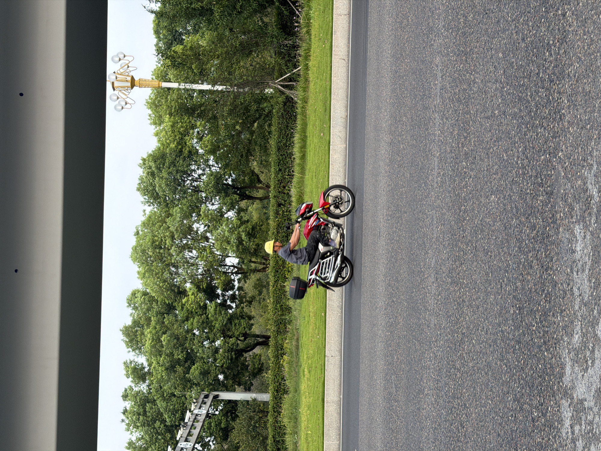 A person wearing a yellow hard hat and a dark grey t-shirt is riding a red motorcycle on a paved road. The motorcycle has a black storage box on the back. The scene is set with a green grassy area and trees in the background. A lamppost with multiple lights is visible to the right of the motorcycle. To the left, a sign structure with some numbers is partially visible. The sky is overcast. The image appears to be taken from a slightly elevated perspective, looking down at the road and the motorcycle. The location is identified as Jiangjin, China.