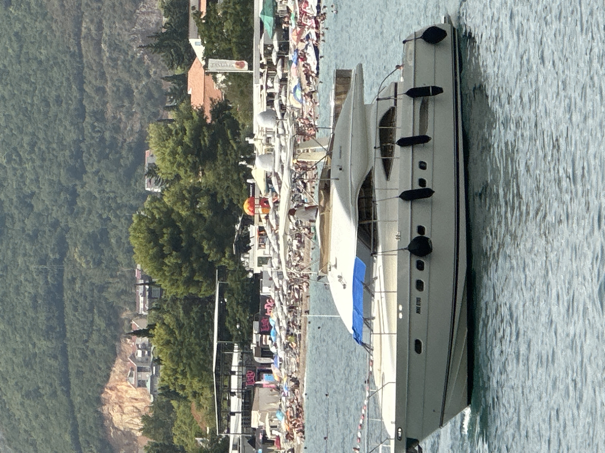 A white motor yacht cruises on the azure waters of the Adriatic Sea, with a crowded beach and a hillside town in the background. The yacht is a prominent subject, featuring multiple decks, dark fenders, and a blue cover over a seating area. A person is visible on the upper deck, looking out towards the sea.
The beach is a hive of activity, with numerous umbrellas, sunbathers, and people enjoying the water. Structures like beach bars and temporary shelters dot the shoreline, suggesting a popular tourist destination. In the distance, the town of Budva, Montenegro, sprawls across a green hillside. Buildings with terracotta roofs and modern structures are nestled amongst lush vegetation, with a rocky, exposed cliff face adding a dramatic element to the landscape. The overall scene depicts a vibrant summer day in a popular coastal resort area.