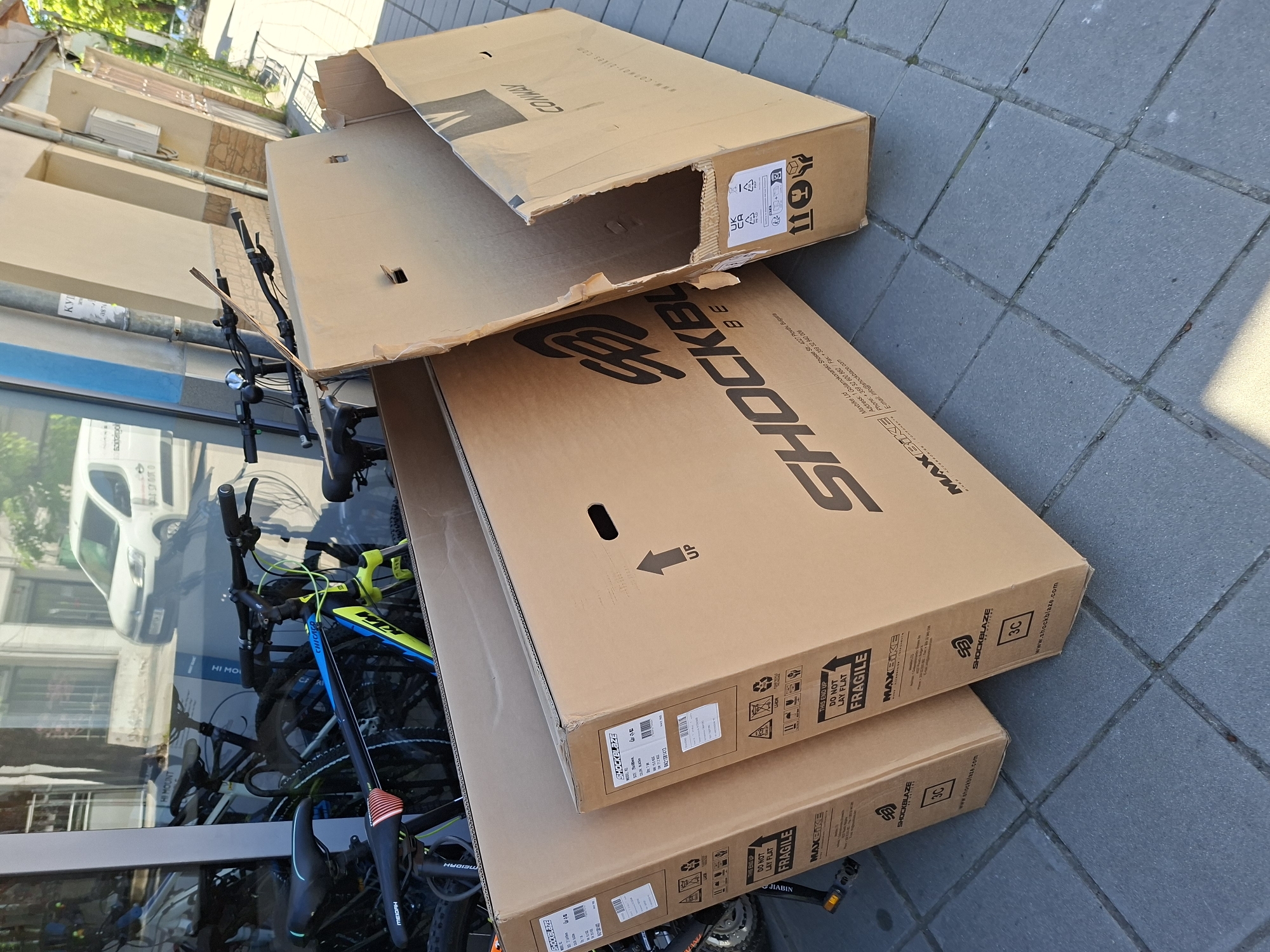 The image shows two bike boxes from "SHOCKBLAZE" and "CONWAY" stacked on a tiled surface, likely a sidewalk or patio. The SHOCKBLAZE box is stacked on top of another SHOCKBLAZE box, while the CONWAY box is partially open, revealing a glimpse of bicycle components inside. Bicycles are visible through a window, suggesting the location is near a bike shop. The address on the box indicates the company is located in Plovdiv, Bulgaria, and the reflections in the window suggest an urban setting, possibly in Gabrovo, Bulgaria. The boxes are marked with "FRAGILE" and "DO NOT LAY FLAT" warnings.