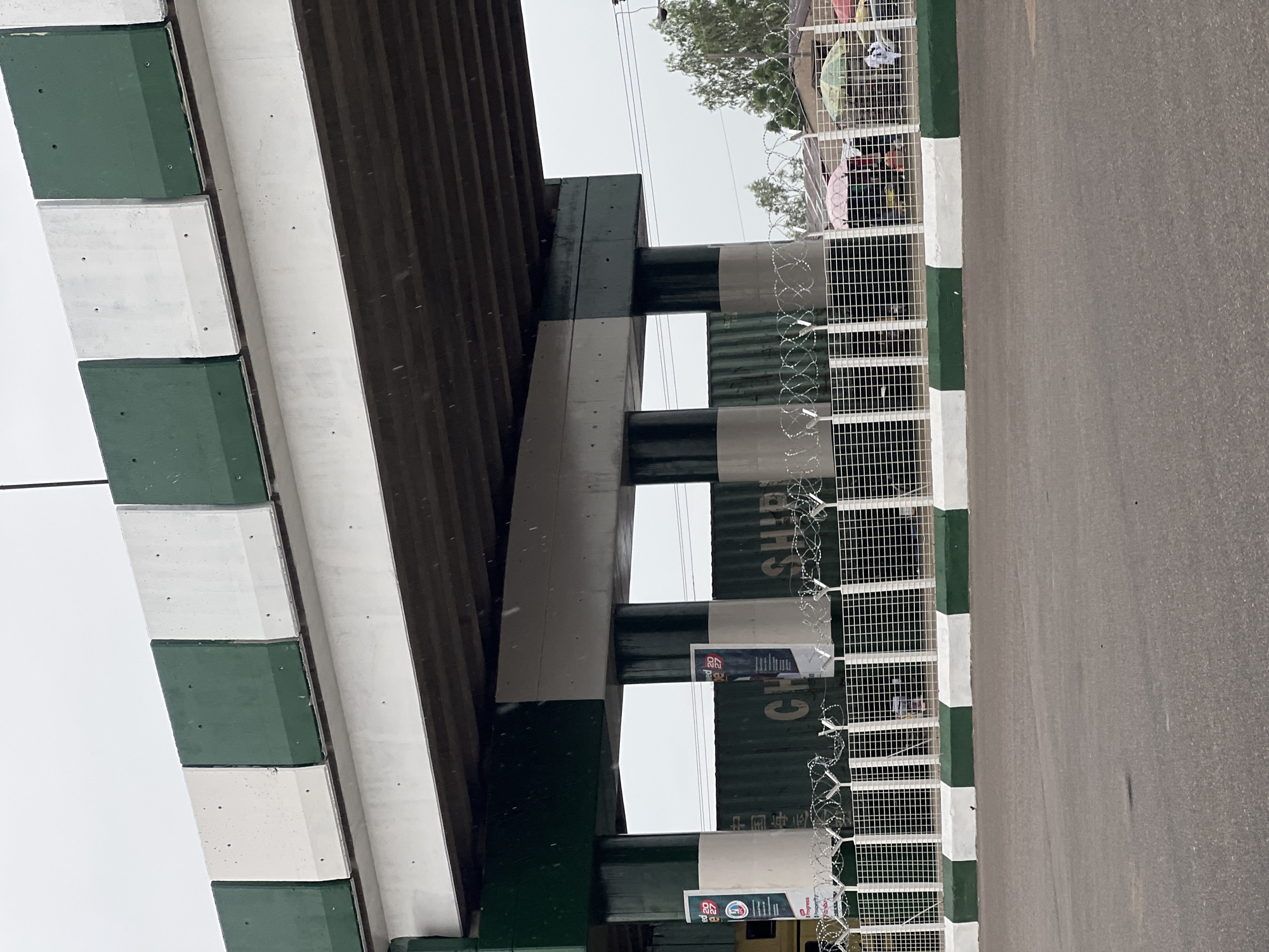 The image depicts a view from a road looking upwards towards an overpass in Jalingo, Nigeria. The overpass is constructed with concrete beams and pillars, painted with alternating white and green sections. Beneath the overpass, a white metal fence with barbed wire topping it runs horizontally. Behind the fence, there are large green shipping containers with visible Chinese characters and the word "SHIP" printed on them. Some vertical banners are attached to the pillars. In the distance, through the fence and containers, several small stalls with umbrellas are visible, with people gathered around them. The sky is overcast and there are visible raindrops, suggesting it might be raining. The road in the foreground is asphalt and has a green and white painted curb.