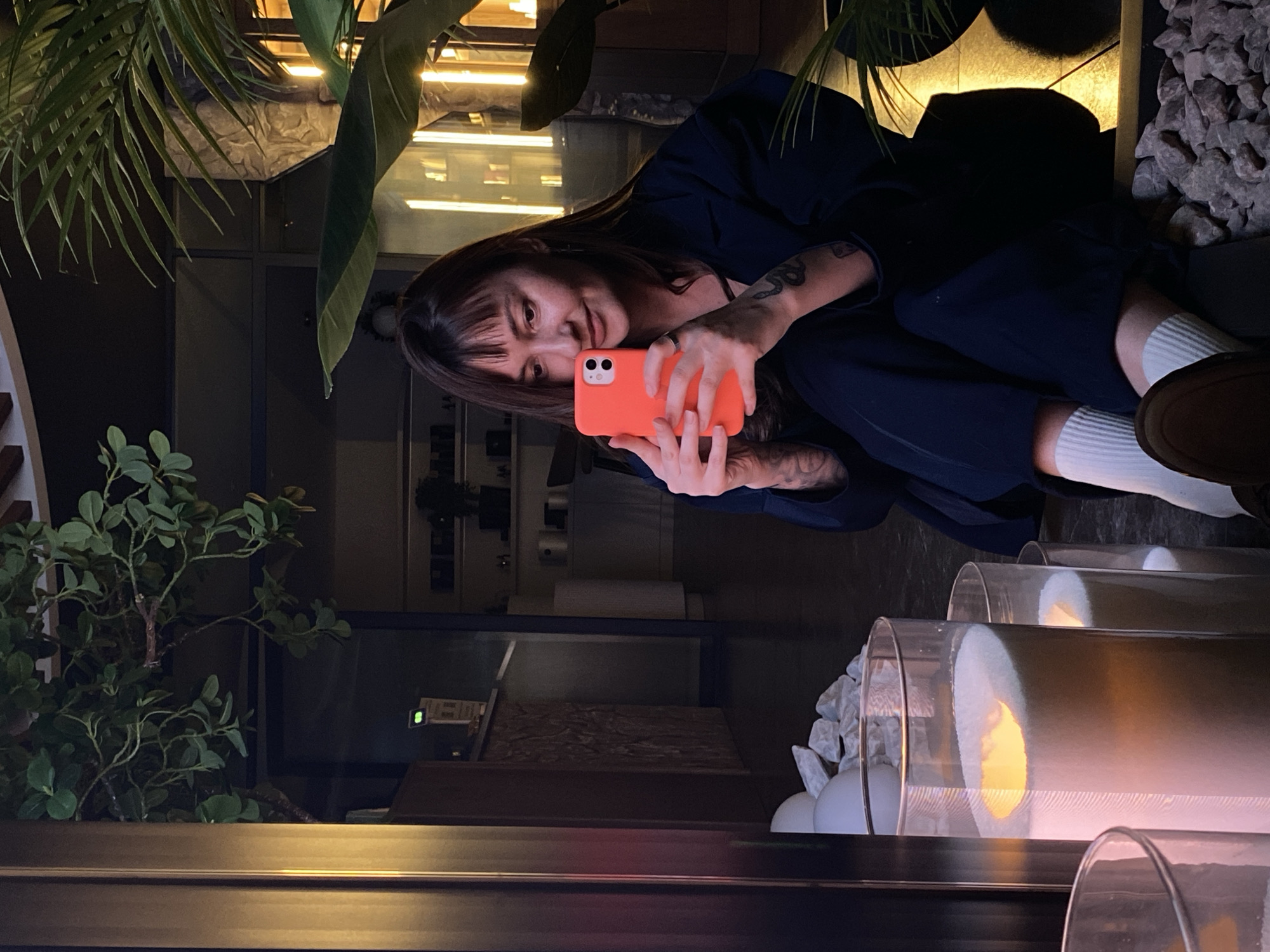 A woman is seated in what appears to be a restaurant or cafe. She is holding an orange smartphone, possibly taking a selfie or using the phone's camera. She is wearing a dark blue or black jacket and white socks. A portion of her footwear, appearing to be brown shoes, is visible. The interior of the building has modern decor, including plants, rocks, and decorative cylindrical lights. The background suggests an urban setting, but the specific location of Votkinsk, Russia, cannot be verified based on the visual information alone.