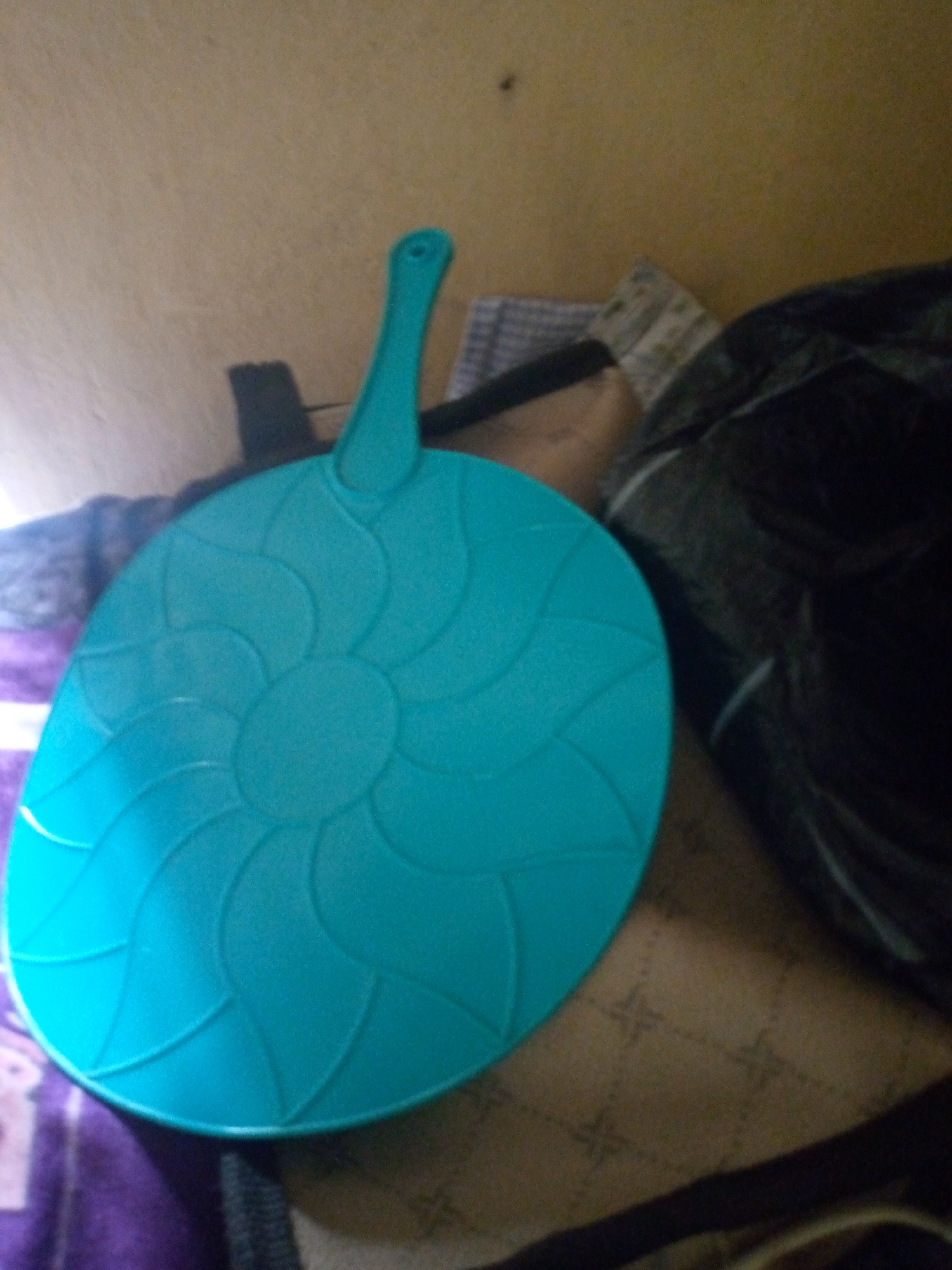 A teal colored hand fan with a petal design is laid on a patterned cloth. The fan has a hole at the end of its handle. The fan is laying on top of a cloth that is brown and white with a geometric pattern. The fan is partially covering another cloth that is purple and white striped. A portion of a black cloth is visible to the right of the fan. The scene appears to be indoors, possibly in a home environment. The lighting suggests it could be daylight, but the shadows are not sharp, hinting at ambient light or possibly artificial light. No people are visible in the frame.