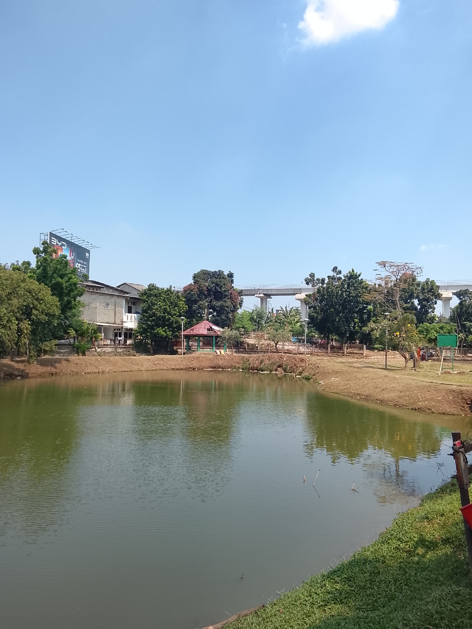 A serene outdoor scene captures a tranquil pond in the city of East Jakarta, Indonesia, on a clear, bright day. The pond, with its calm, green waters, reflects the surrounding environment, creating a mirror-like effect. A lush green embankment lines the edge of the pond, with well-maintained grass adding to the natural beauty of the area. In the background, a cluster of trees stands tall, providing shade and a touch of wilderness. A red-roofed gazebo offers a picturesque resting spot, while a multi-story building, possibly a residential or commercial structure, is visible in the distance. A large billboard, advertising a product or service, adds a modern touch to the otherwise natural landscape. The scene is further enhanced by an elevated bridge, a testament to the city's infrastructure. The overall ambiance is one of peace and serenity, a perfect escape from the hustle and bustle of city life.