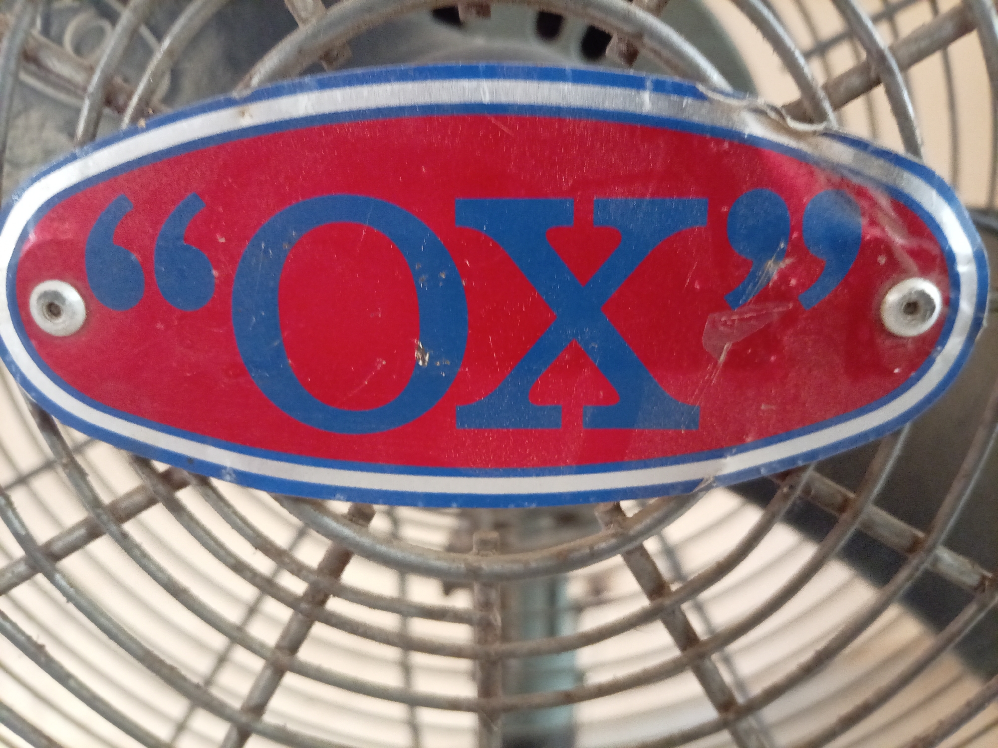 The image shows the close-up of the front grille of a fan. The grille is metallic, with a visible circular pattern. Centered on the grille is an oval badge, red in color, with a blue border and white line separating the red from the blue. The word "OX" is written on the badge in blue, flanked by quotation marks. The badge is attached to the grille with screws in each of its ends. The fan appears to be in used condition, exhibiting wear and tear, and some dirt or discoloration. There is no indication of Orie Uratta, Nigeria, or people in this image.