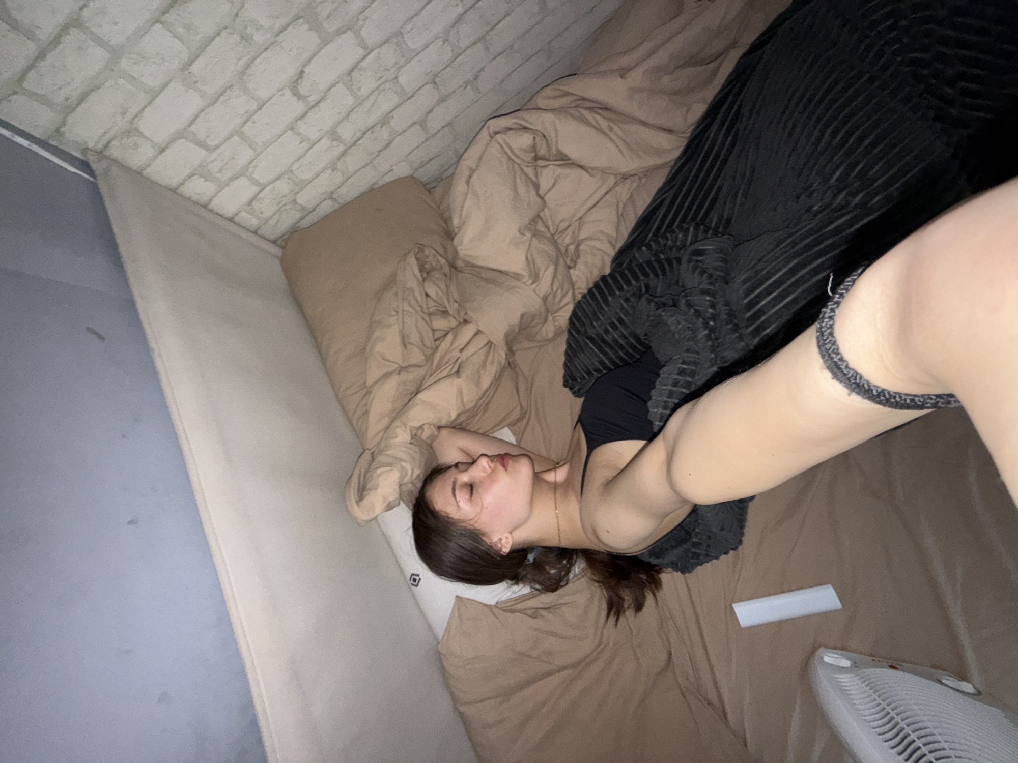 A photograph depicts a woman lying in a bed in a room with light-colored brick wallpaper and light-colored sheets. She has dark hair and is wearing a dark top. Part of her arm is visible, as is a small fan and a white rectangular object. The location in Sofia, Bulgaria, is unknown.