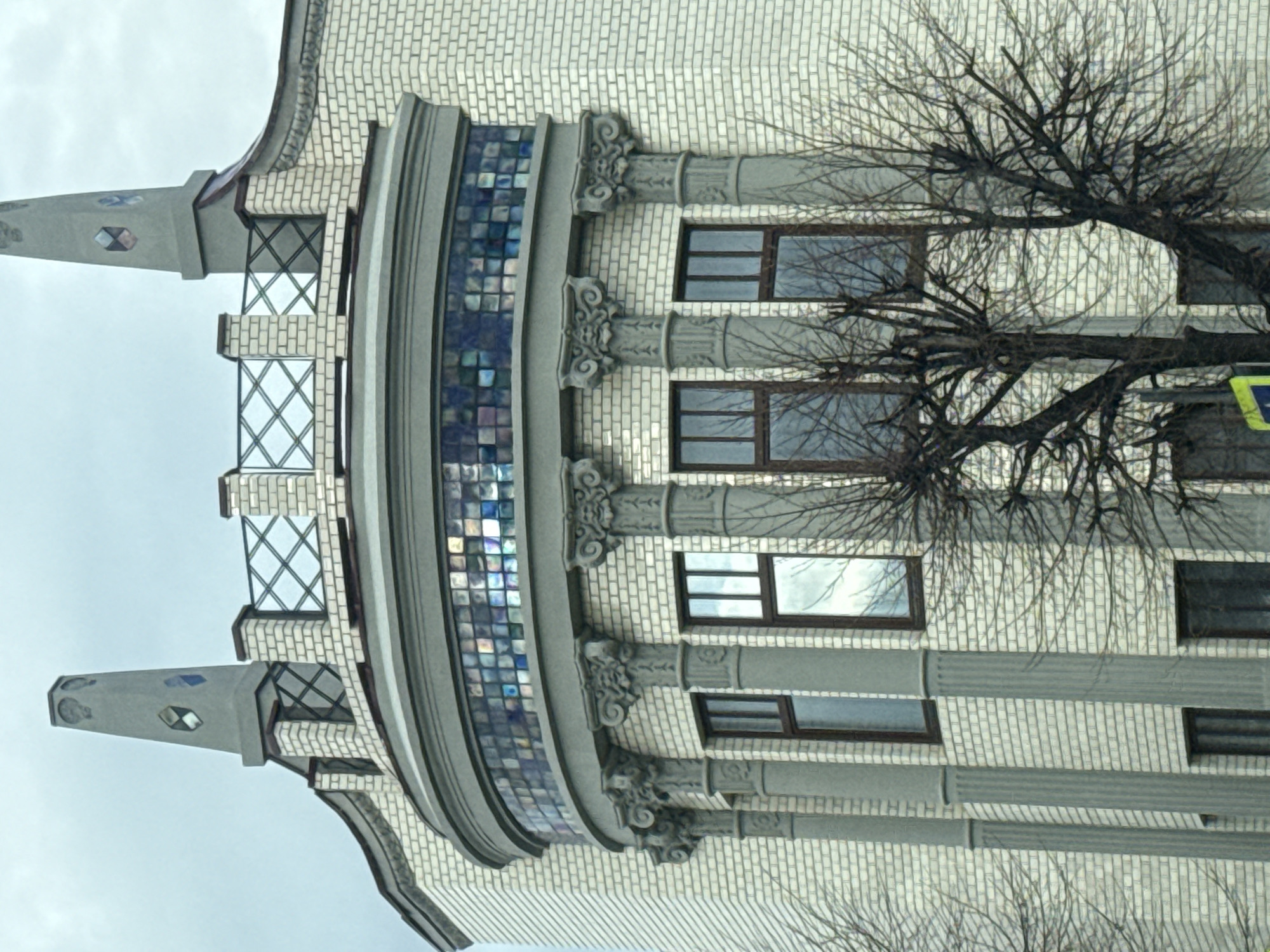 Here is a description of the image:
The image depicts a building in Syzran, Russia, showcasing architectural details. The structure is characterized by light-colored brickwork, adorned with decorative elements such as ornate columns and a mosaic-tiled curved section. Several windows with dark frames are visible on the facade. The building's corner features distinctive spire-like projections with decorative diamond shapes. A bare tree obscures parts of the building, its branches reaching across the facade. The sky appears overcast, suggesting a dull day.