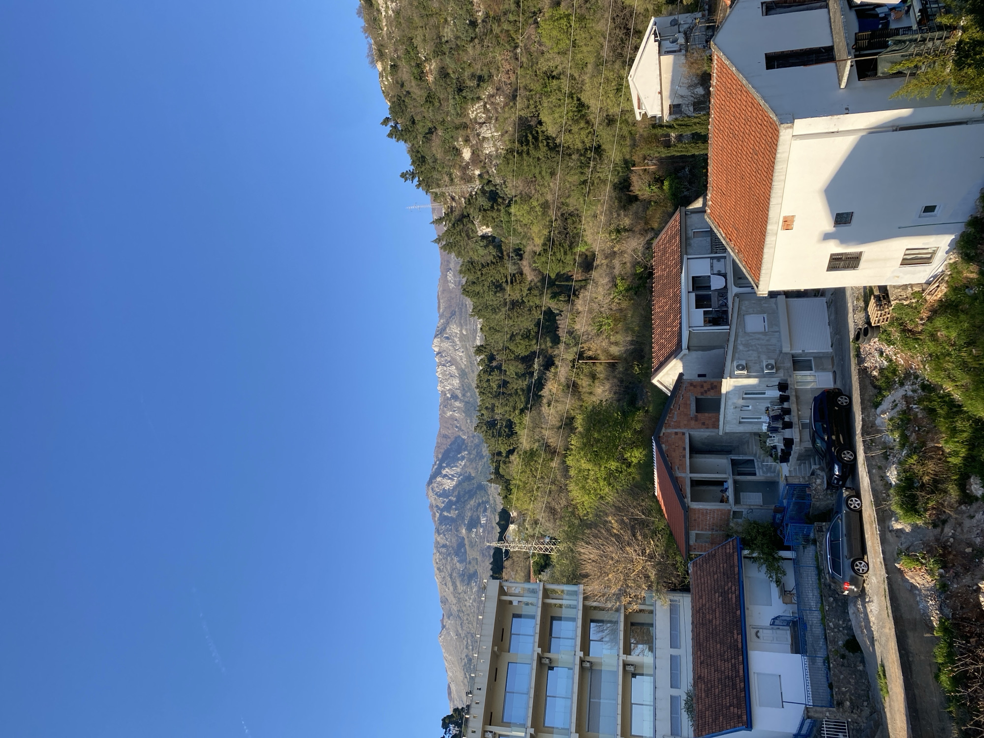 The image depicts a view in Budva, Montenegro. The scene features a mountain range on the left side of the frame, transitioning to a mix of vegetation and residential buildings on the right. The sky above is clear and blue. Buildings include multi-story structures with glass facades and houses with red-tiled roofs. Cars are parked along a narrow road. Power lines are visible against the mountain backdrop. The image is oriented vertically.