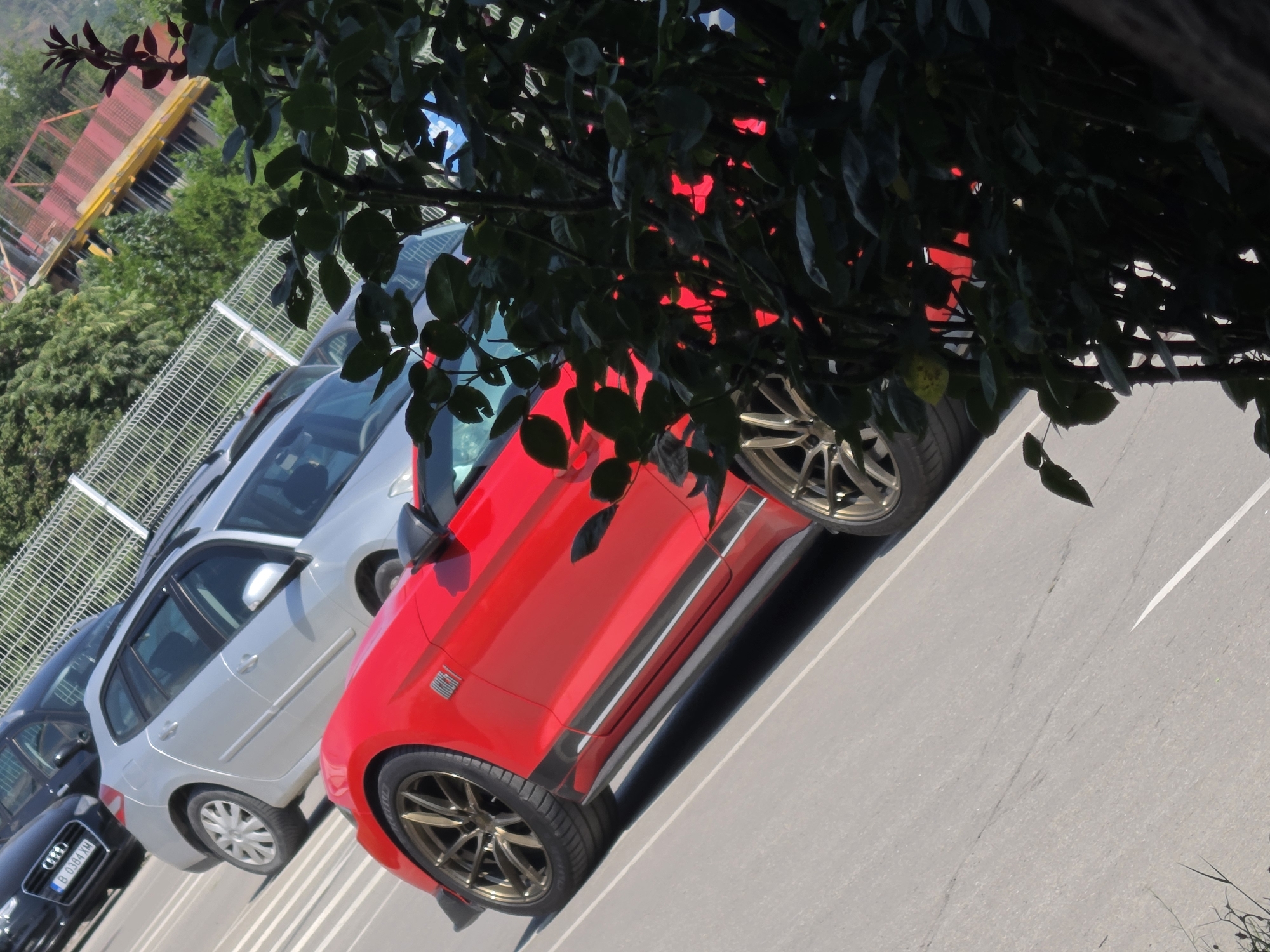 The image depicts a street scene, likely in Varna, Bulgaria, given the context. In the foreground, a bright red sports car, possibly a Ford Mustang, is parked on an asphalt road. The car is angled, with its front wheel and part of its side visible. The wheel features a bronze-colored rim. Behind the red car, a silver station wagon is parked, and further back, a dark-colored sedan is visible. The cars are aligned in what appears to be a parking area or along a street. A metal fence runs along the background, separating the road from a lush green area with trees. In the upper right portion of the image, a section of a yellow and red structure, possibly part of a playground or construction, is visible. The overall lighting suggests it is daytime, with bright sunlight casting shadows. No people are visible in the image. The scene is static, with the vehicles appearing to be stationary.