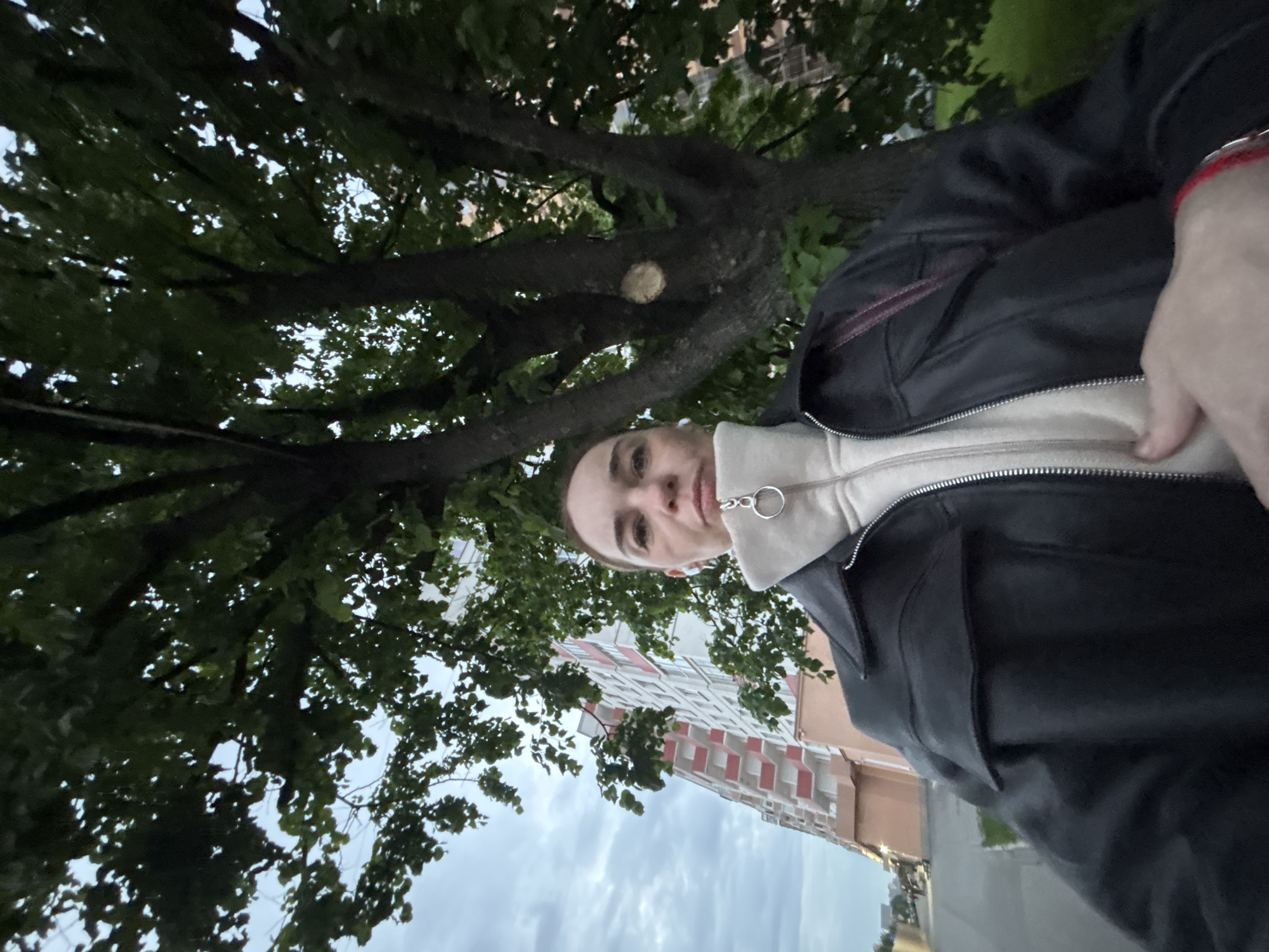 The image depicts a person wearing a black leather jacket over a light-colored hooded sweatshirt. The individual is standing beneath a tree with dense green foliage. A building with red and white patterns is visible in the background. The sky is overcast. The location appears to be an urban area in the city of Unknown, Unknown.