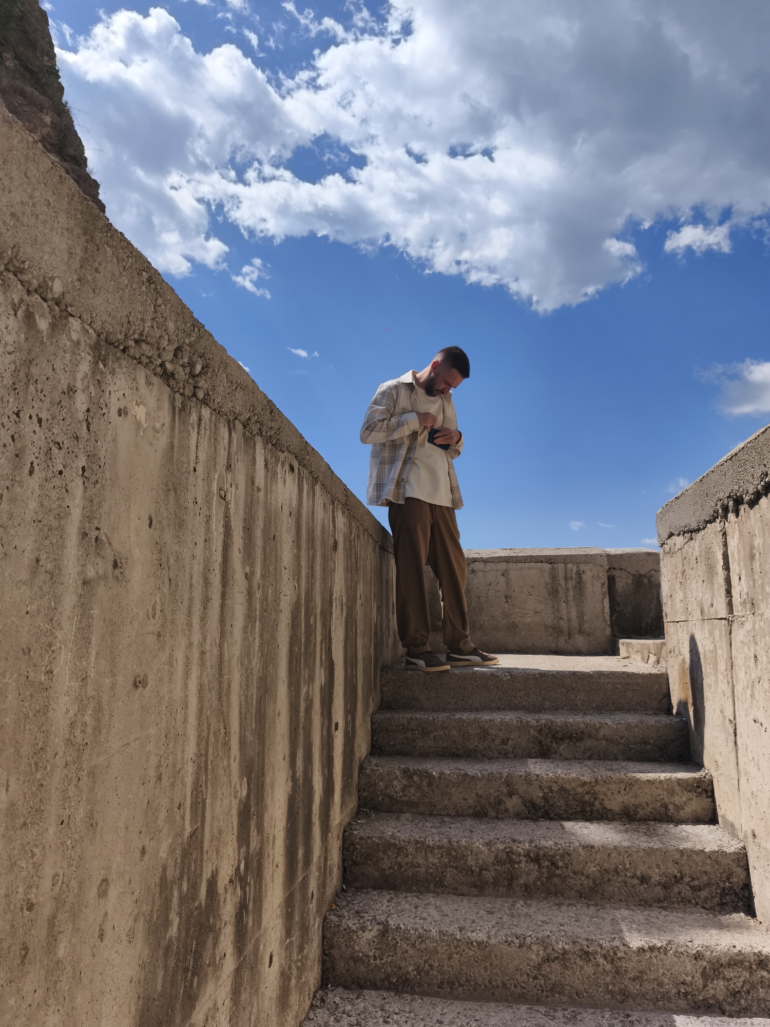A man stands on stone steps enclosed by concrete walls in Rafailovići, Montenegro, under a partly cloudy sky. The man is fair-skinned, with short brown hair and a beard. He is wearing a white t-shirt, a tan plaid overshirt, brown trousers, and two-toned sneakers. He is looking down at a small, dark object in his hands, likely a phone or wallet. The walls are made of rough, textured concrete, stained with weathering. The steps are also concrete. The sky is blue with scattered white clouds. The scene suggests an outdoor location in a historical or architectural setting.