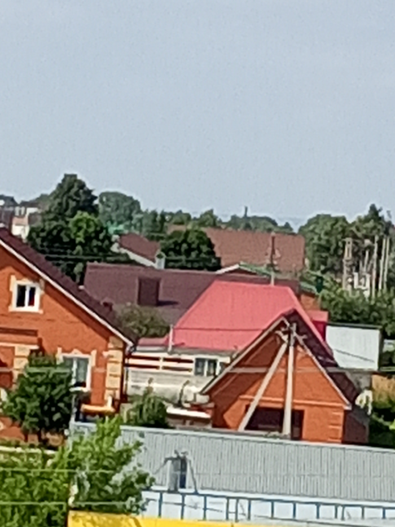 The image shows a cluster of residential buildings under a clear sky. The foreground is dominated by the roof of a grey, corrugated structure, likely a fence or a large shed, with blue detailing along its top edge. Behind this, a bright yellow horizontal element is partially visible at the bottom.
The middle ground features several houses with distinct architectural styles and roofing materials. To the left, a brick house with a red-brown roof has a white-framed window visible on its upper story. Adjacent to it, another building has a red-brown roof and a lighter colored wall. Further to the right, a prominent house stands out with a bright red, gabled roof. This structure has exposed wooden beams forming a triangular frame on its front facade, suggesting it might be undergoing construction or renovation, or is designed in a rustic style. The walls of this house appear to be orange brick. More buildings with red-brown roofs are visible in the background, interspersed with lush green trees.
The background is filled with dense foliage and more buildings, suggesting a suburban or semi-rural setting. The overall impression is one of a residential area, possibly a village or a district on the outskirts of a city. The lighting indicates it is daytime, likely sunny, given the bright colors and clear sky.