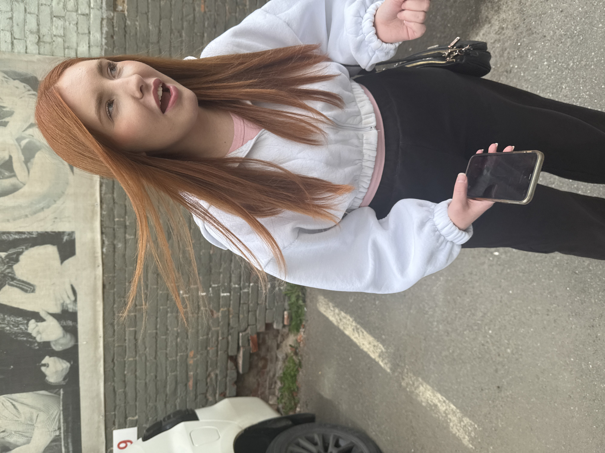 A young woman with long, red hair stands on a paved surface in front of a brick wall. She is wearing a white cropped zip-up hoodie over a pink t-shirt and black pants. She is holding a smartphone in her left hand and her right hand is open, palm up. A black purse with gold accents is slung over her right shoulder. To her left, a white vehicle is partially visible, with a sign showing the number "9" attached to its side. The background features a mural or large black and white photograph on a lighter-colored surface above the brick wall. The scene appears to be an outdoor location, possibly a parking lot or street in Perm, Russia, given the context provided.