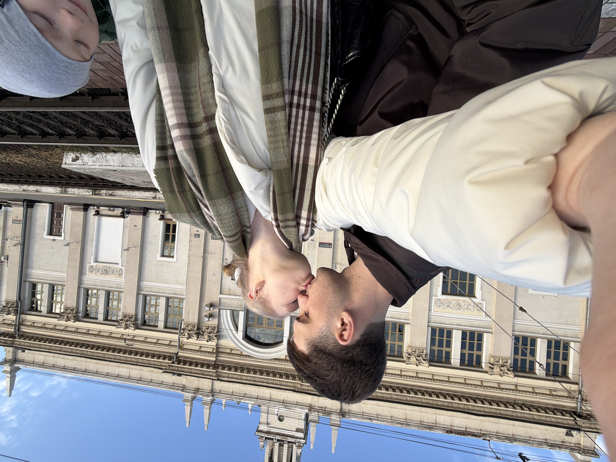 A couple is kissing in front of a building in Krasnodar, Russia. The woman has blonde hair and is wearing a white jacket and a plaid scarf. The man has dark hair and is wearing a dark jacket. The building is a light color and has many windows. The sky is blue. In the upper left corner, a child can be seen wearing a grey beanie and what appears to be a white jacket. They appear to be standing on a platform with metal barriers.