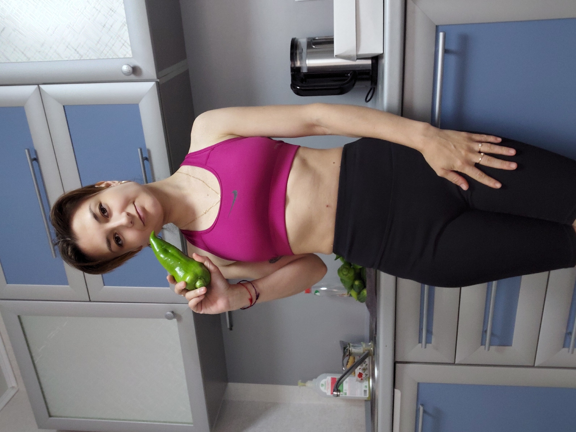 A woman is standing in a kitchen, holding a green pepper to her face. She is wearing a pink sports bra and black leggings. The kitchen has grey cabinets with blue accents. There is a sink with a bottle of soap to the left of the woman, and a coffee maker and some boxes to her right. The overall impression is one of a casual, domestic setting.