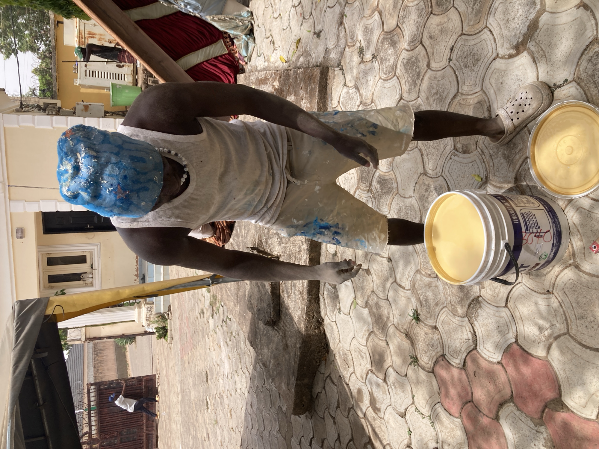 A person wearing a blue hair covering, a white tank top, and shorts with paint splatters stands outdoors on a paved surface in Jos, Nigeria. The person is near two paint buckets, one open and filled with yellow paint and the other with its lid nearby. They are wearing white clogs. The surrounding area includes buildings, a partially visible vehicle or structure with a yellow tarp, and other people in the background, suggesting an urban or residential setting. The paving stones are interlocking and appear aged, with some discoloration.