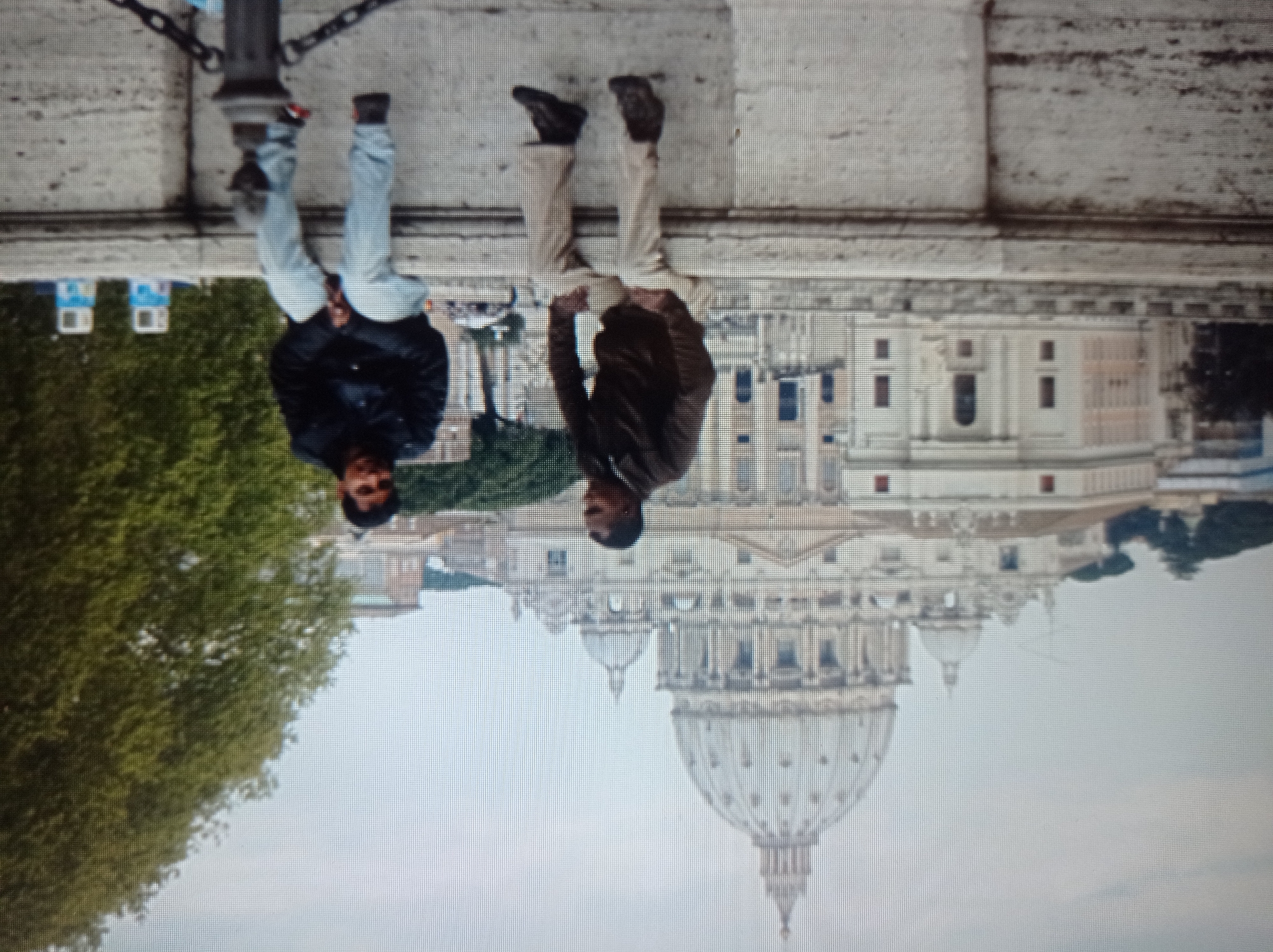 The image depicts two individuals seated on a stone wall with St. Peter's Basilica in Vatican City visible in the background. The person on the left wears a brown jacket and beige pants, while the person on the right is dressed in a black jacket and light blue jeans. The sky is overcast. The image is pixelated, affecting clarity and detail. The reference to Mukachevo, Ukraine is extraneous to the visual content of the image.