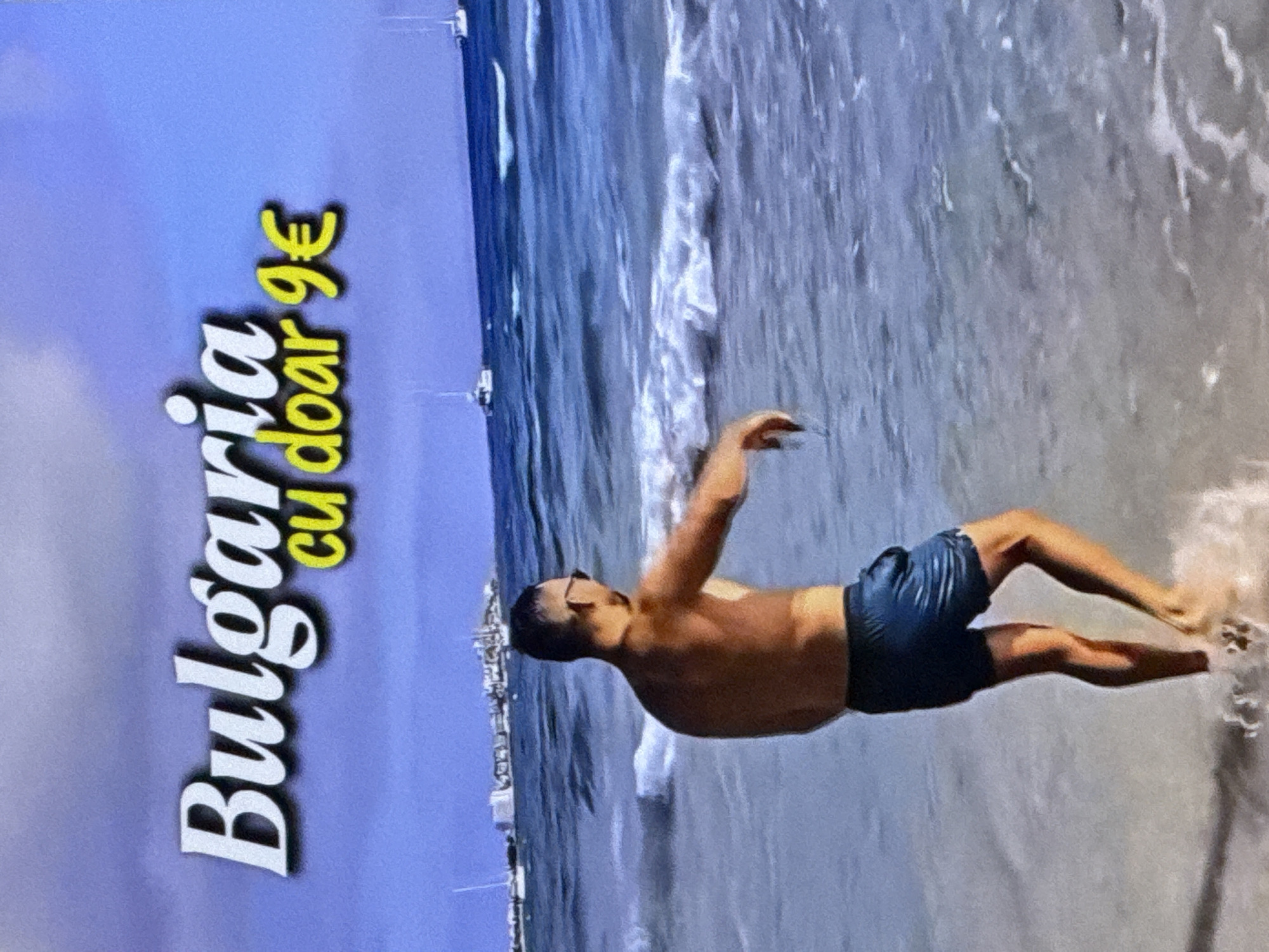 This media file features a man in dark blue swim trunks standing in shallow water at a beach. The water is breaking around his legs, and he appears to be in motion, with his arms raised. He is wearing glasses and has a tanned complexion. In the background, there is a body of water with visible waves and a hazy shoreline with structures that could be buildings or boats. The sky is a light, hazy blue.
To the left of the image, text is overlaid on a purple background. The text reads "Bulgaria cu doar 9€". This suggests that the image is related to a travel advertisement for Bulgaria, with a price of 9 Euros. The overall impression is of a sunny day at a beach, potentially promoting an affordable trip to Bulgaria. The man's pose and the waves suggest a dynamic and refreshing atmosphere.
