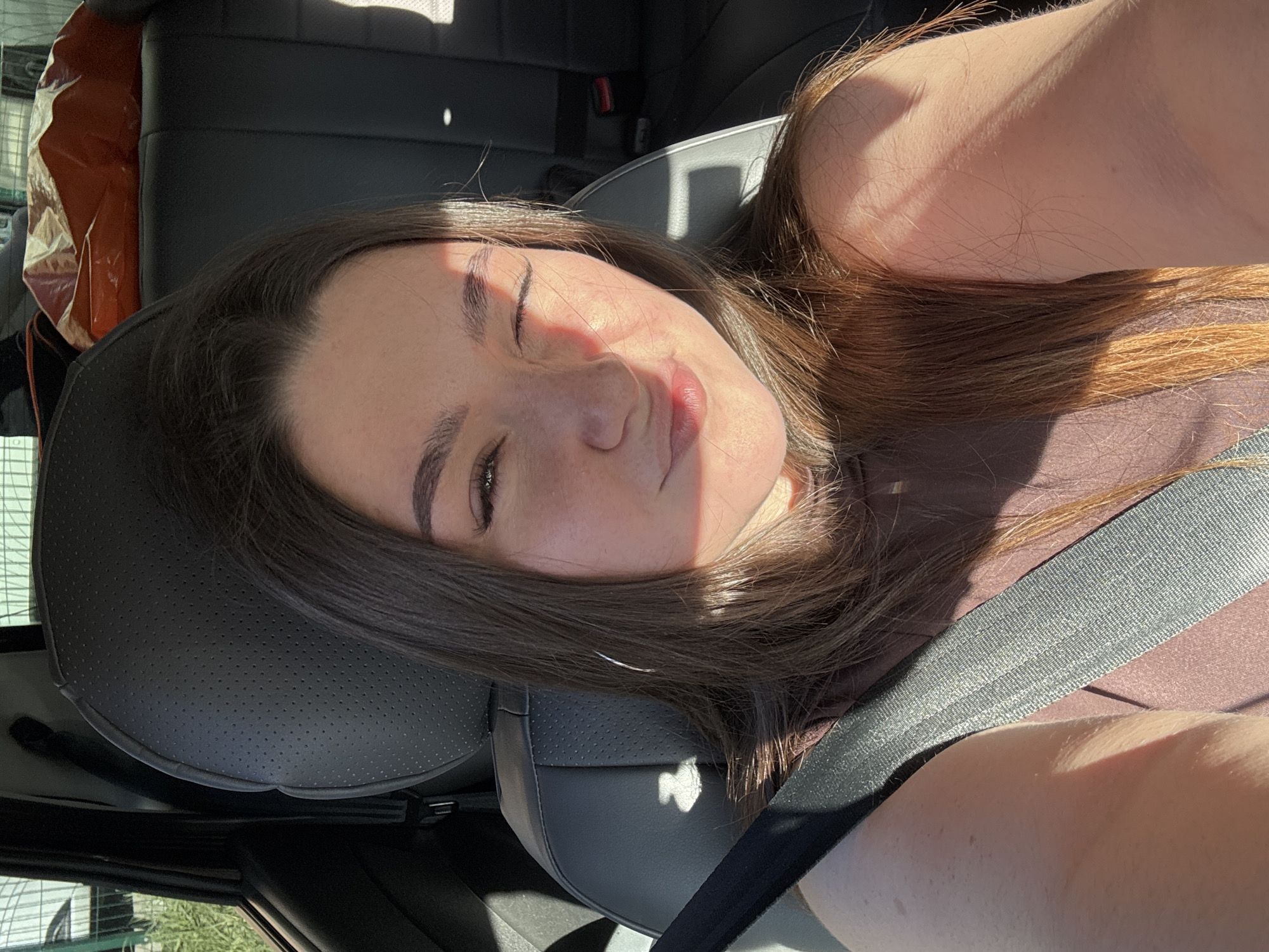 A woman is seated in the driver's seat of a car, wearing a seatbelt and a brown top. Her head is resting on the headrest. She has brown hair and her eyes are partially closed, seemingly squinting in the sunlight. The interior of the car is visible, including the black leather seats and a glimpse of the car's interior behind her. The setting is not identifiable as Perm, Russia from the visual information.