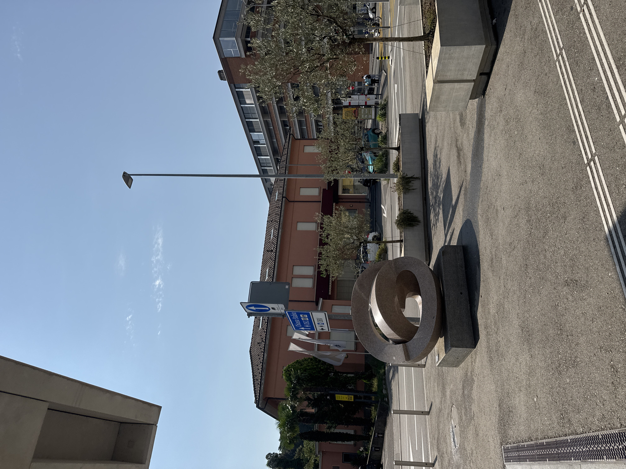 This is a daytime, eye-level, outdoor, full shot of a street scene in Mendrisio, Switzerland. The primary focus is on a large, abstract stone sculpture in the foreground. The sculpture is composed of interlocking, spiraling forms and rests on a dark granite base. Behind the sculpture, a coral-colored building with a tiled roof is visible. To the right of this building, a modern, multi-story brick building with large windows stands. In front of the buildings, there are trees, planters, streetlights, and road markings. A blue and white street sign indicating "Chiasso Stazione" and "OBV" is also present. Several cars, a motorcycle, and a few pedestrians are visible in the background on the street and sidewalk. The sky is clear and blue.