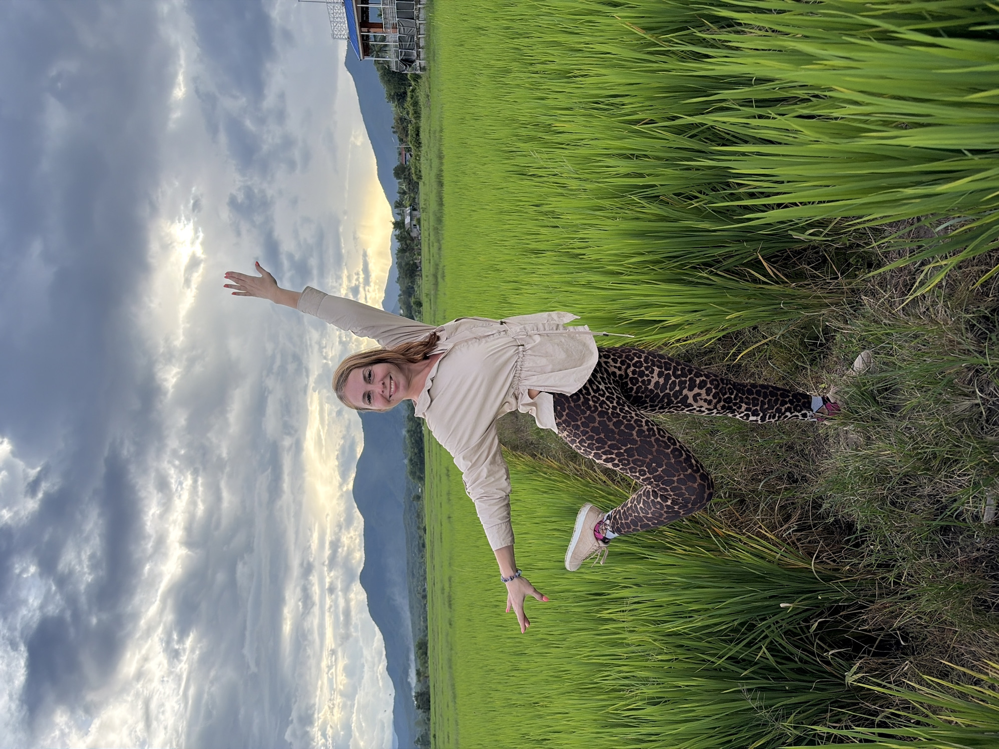 In Ban Pong, Thailand, a woman stands in a lush green rice field under a cloudy sky. She is posing with her left leg bent at the knee and extended backward, while her right arm is raised and her left arm is extended to the side. The woman is wearing a tan collared shirt with cinched waist and leopard-print leggings. The background shows a mountain range and a building with a blue roof. The rice paddy is dense with vibrant green stalks, and a narrow dirt path is visible where the woman is standing.