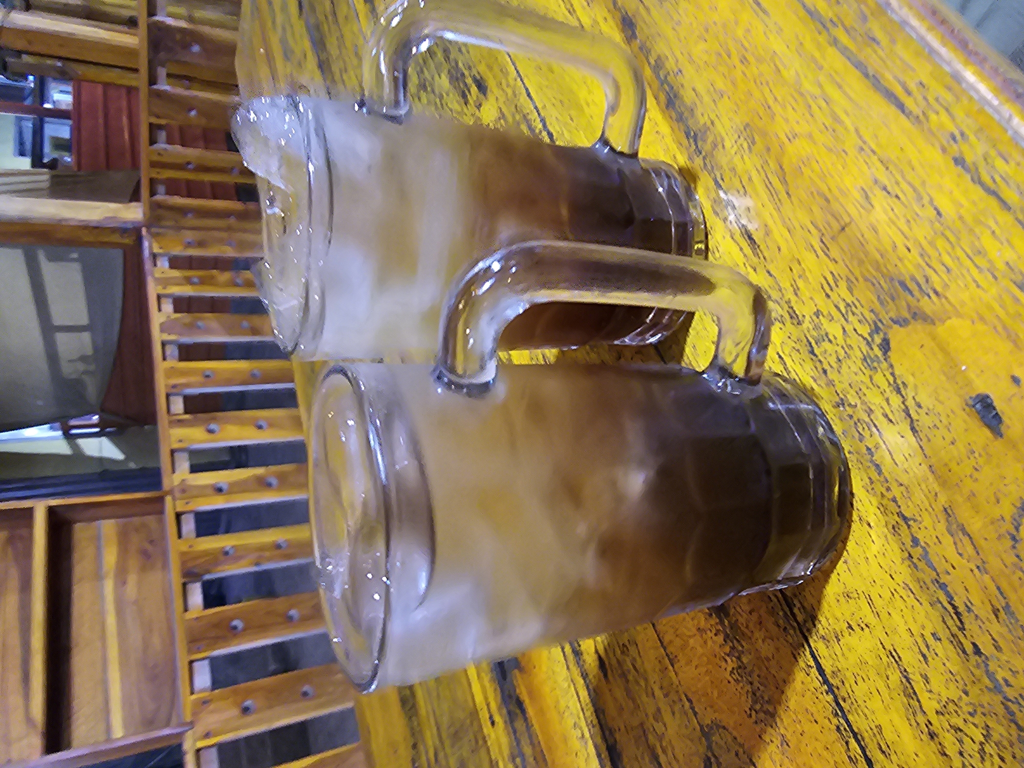 Two glasses of iced tea with ice cubes sit on a yellow wooden table. The glasses are tall and clear, with thick handles. The liquid inside is a deep brown color with a lighter, foamy layer at the top. In the background, there is a wooden slatted bench and a wooden structure that appears to be part of a building or furniture. The lighting is dim, suggesting an indoor or shaded outdoor setting. The image does not contain enough information to determine the specific location in Jenu, Indonesia.