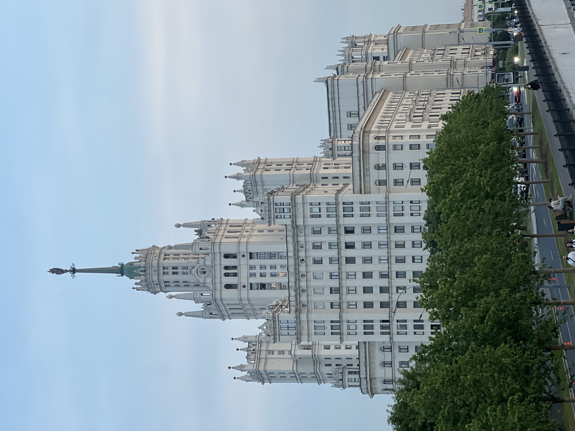The image captures a grand, ornate white skyscraper with a spire, characteristic of Stalinist architecture, dominating the skyline. The building features numerous windows, decorative elements, and smaller turrets. In the foreground, a row of lush green trees lines a sidewalk with a black railing. Beyond the trees, a street with several cars and a few pedestrians is visible. The sky is a pale, cloudy blue, suggesting it is either early morning or late evening. The overall scene conveys a sense of urban grandeur and a peaceful, possibly early evening, atmosphere.