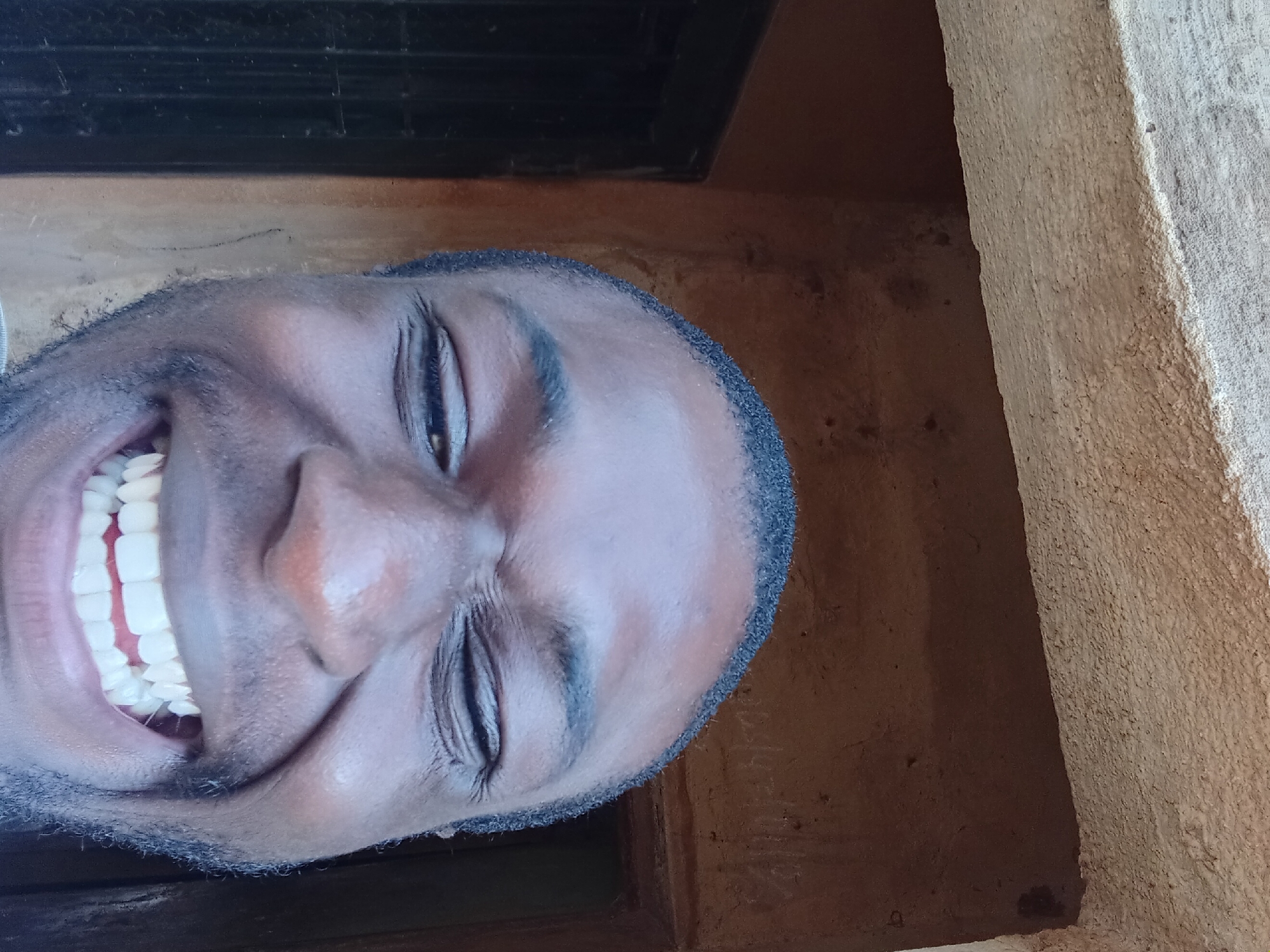 A close-up, low-angle selfie shows a Black man's face filling most of the frame. He is smiling broadly, with his eyes squinted shut and his teeth showing. The man is wearing a grey t-shirt. He appears to be standing outdoors, with a textured brown wall and a rough, light-colored overhang visible behind him. A dark doorway is partially visible on the left side of the frame. The scene is likely in Bukuru, Nigeria, given the context.