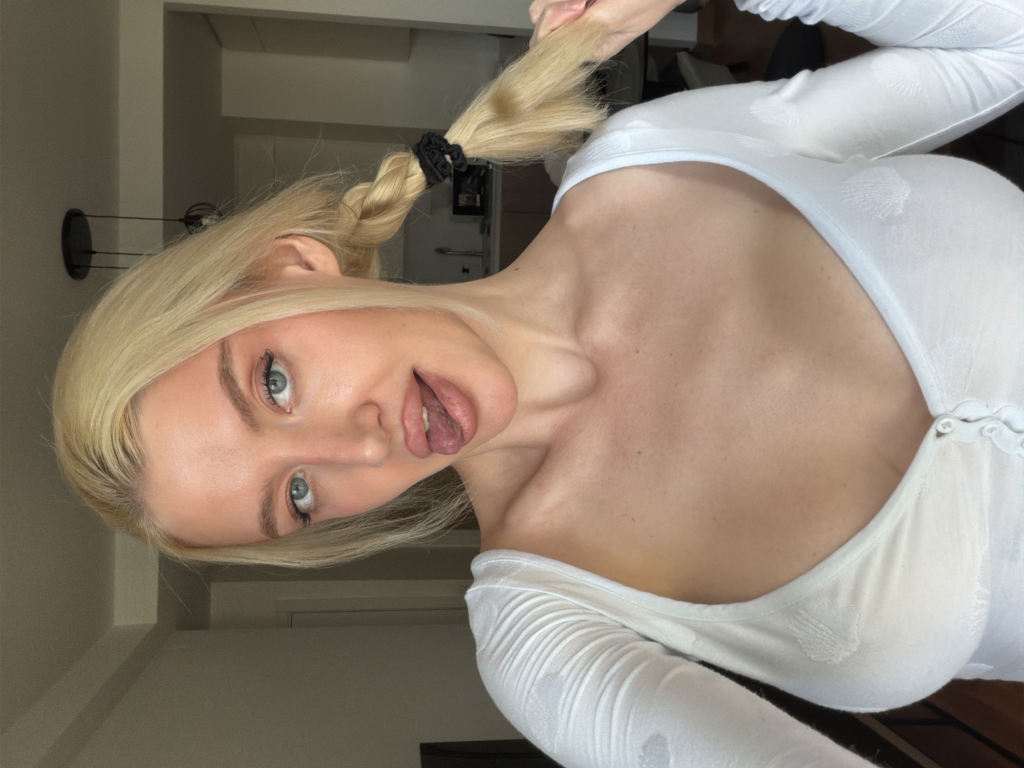 A woman with blonde hair in a ponytail is positioned at an angle, wearing a white long-sleeved shirt with buttons. She is sticking her tongue out. The background includes a door, a light fixture, and a surface, possibly a counter or shelf. The location cannot be determined from this image.