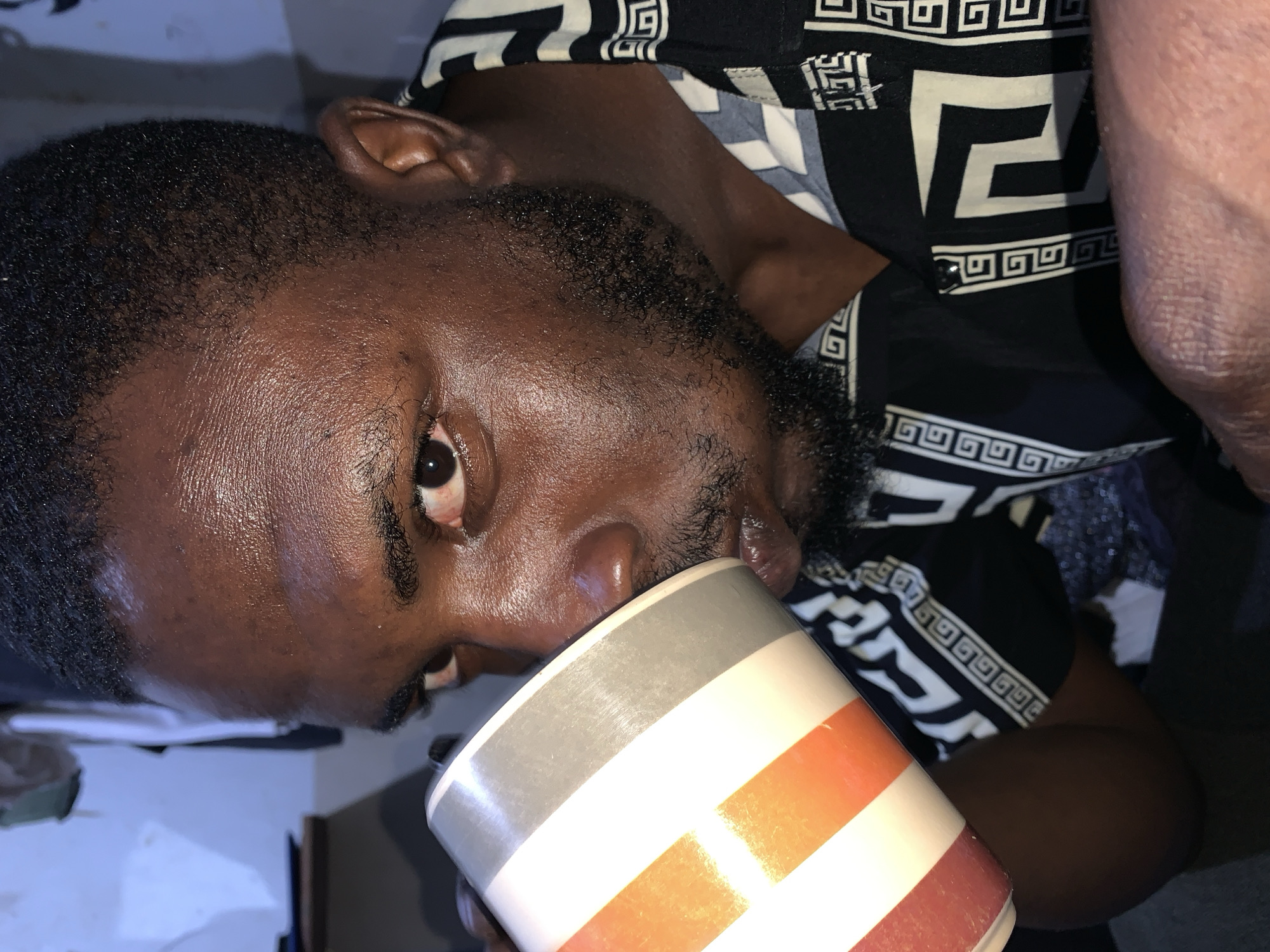 In the city of Rafin Par, Nigeria, a close-up shot captures a man drinking from a mug. The man, with dark skin and a short beard, is wearing a black and white patterned shirt. He is holding a mug with white, orange, and gray stripes to his lips, mid-drink. The background is out of focus, but suggests an indoor setting with a light-colored wall and some objects on a surface. The lighting is artificial, creating highlights on the man's face.