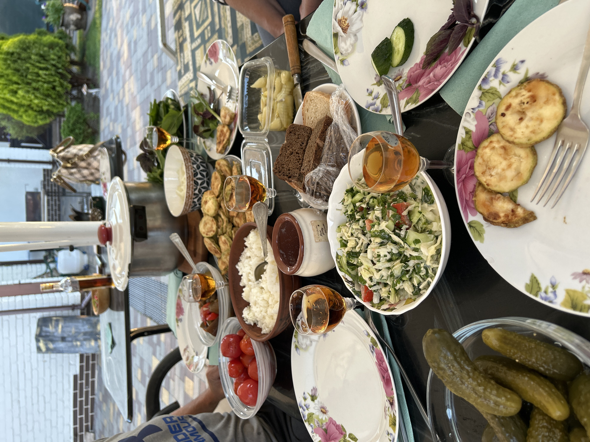 An outdoor table is laden with a variety of dishes, suggesting a meal setting, possibly a picnic or al fresco dining. The table is covered with a patterned tablecloth and set with plates, bowls, and glasses. Among the food items are a salad, rice, tomatoes, fried zucchini, bread, and pickles. There are also glasses filled with an amber liquid, likely wine or juice. A large pot with a lid is visible on the table, and a bottle of what appears to be cooking oil or liqueur stands nearby. In the background, there's a brick wall, a paved patio, and lush greenery, including a prominent bush. The scene is illuminated by natural daylight.