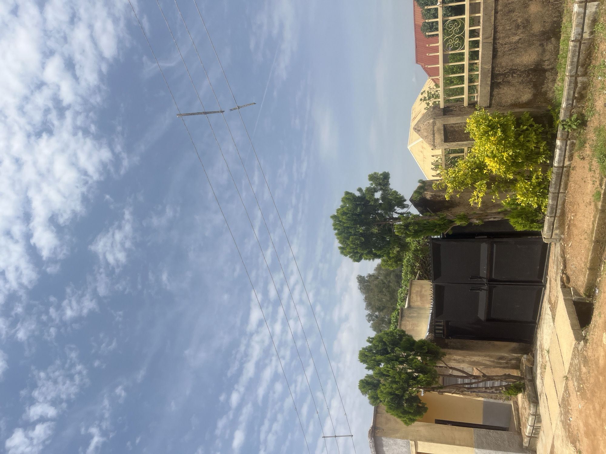 The image shows a residential property in Gura Top, Nigeria, on a partly cloudy day. In the foreground, a dirt road leads to a large, black metal gate, flanked by a building with a yellow and gray facade. Several evergreen trees are planted on either side of the gate. Behind the gate, a concrete wall topped with a decorative metal fence encloses the property. Further in the background, the roofs of houses are visible, including one with a yellow, conical structure and another with a red roof. Power lines stretch across the sky, with one pole appearing broken. The sky is filled with scattered white clouds against a pale blue backdrop. There are no people visible in the image.