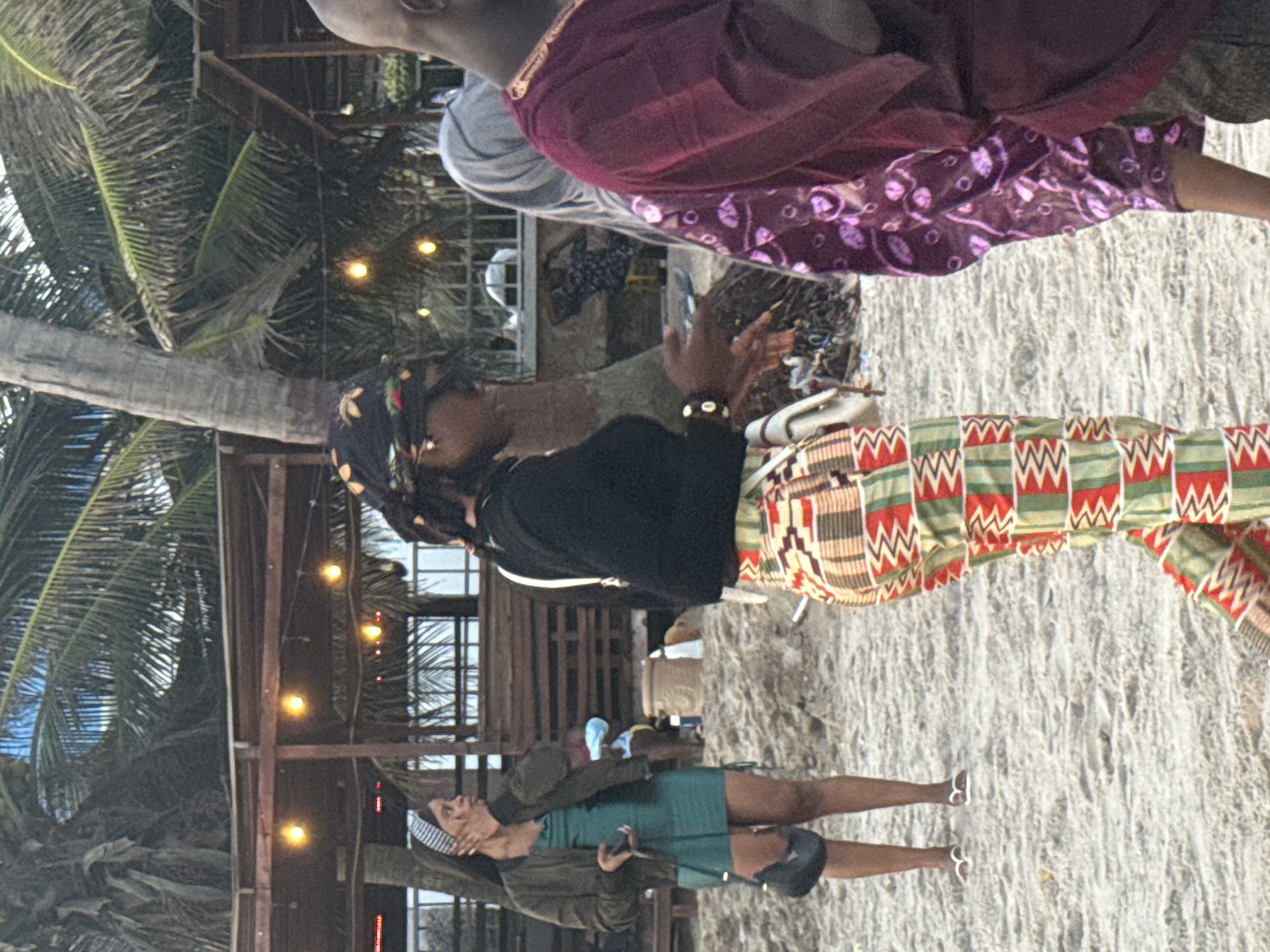 The photograph shows a beach scene in Mopo Onijebu, Nigeria. The sandy beach forms the ground, with a palm tree on the left side and a wooden structure adorned with string lights in the background. Three people are visible. The closest person wears a green dress and jacket, and has a headband. The next wears a black top and patterned pants, with a head wrap. The last person wears a purple shirt with patterned sleeves.